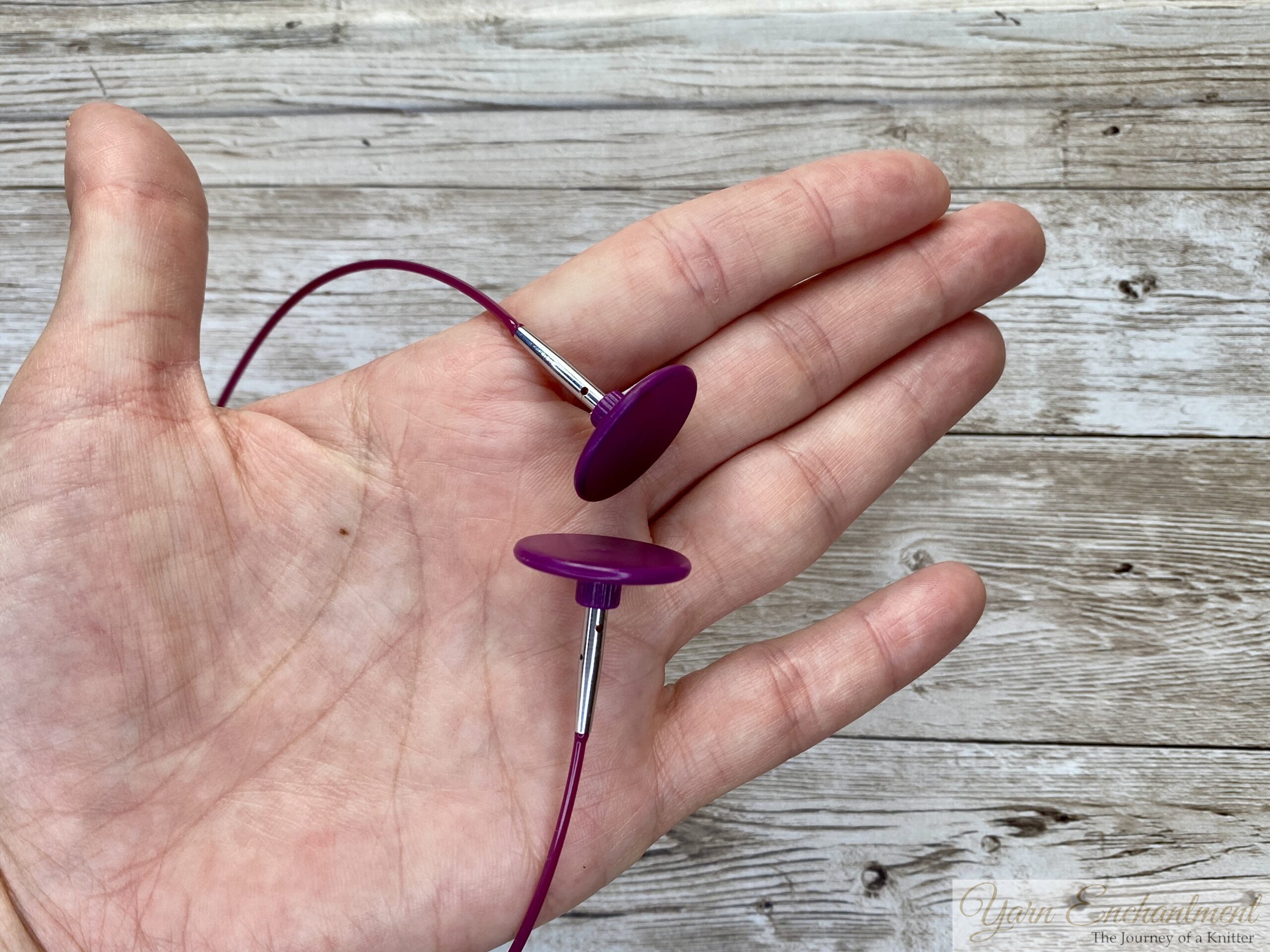 A close-up of a hand holding a pink cable with two purple stoppers attached, used for securing stitches during knitting.