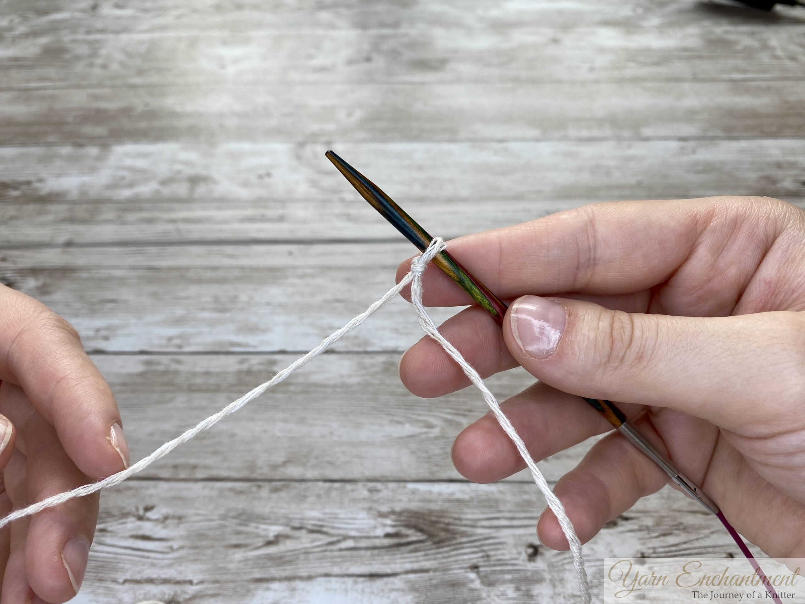 Two hands holding knitting needles and white yarn. The wooden needle is being prepared with a slip knot, indicating the start of a provisional cast on