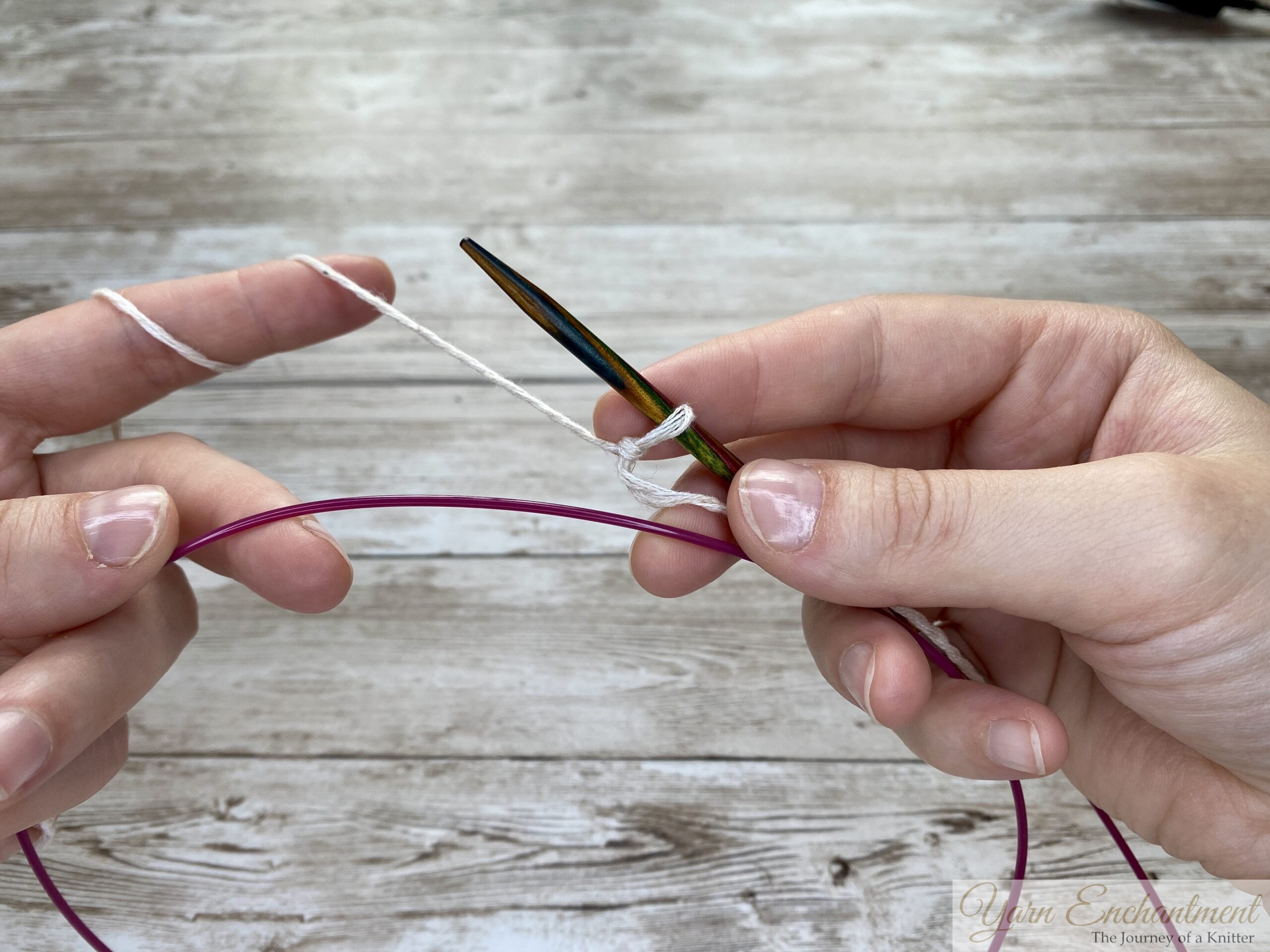 Two hands demonstrating the provisional cast-on technique. The wooden knitting needle holds a few loops of white yarn, while the pink cable is positioned below to act as the spare holder for the cast-on stitches