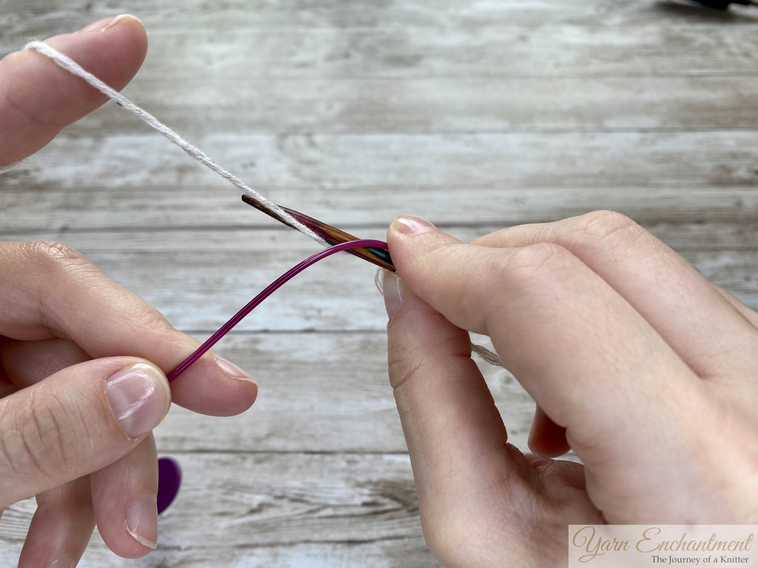 Two hands demonstrating the provisional cast-on technique. The wooden knitting needle holds a few loops of white yarn, while the pink cable is positioned below to act as the spare holder for the cast-on stitches