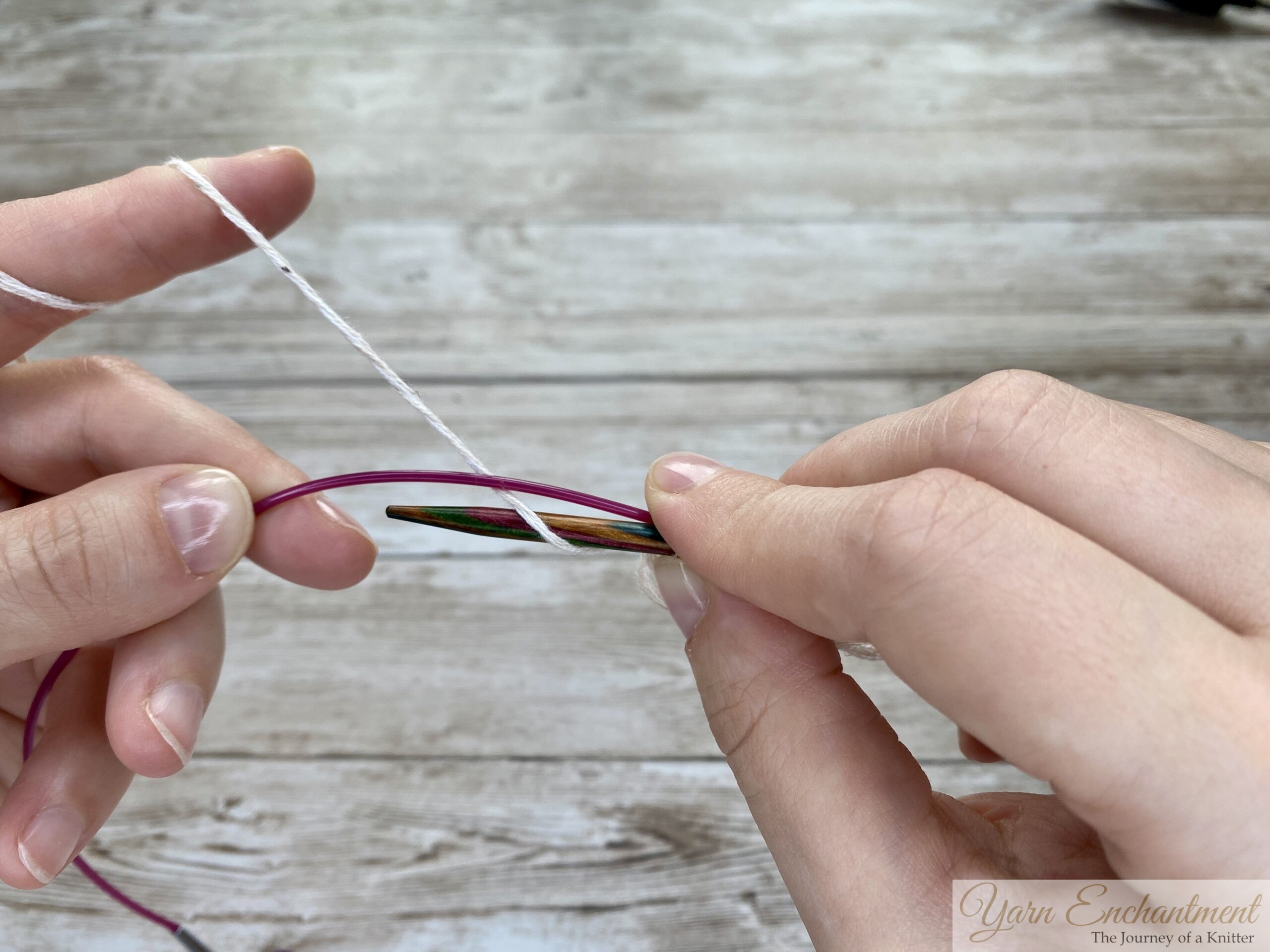 Two hands demonstrating the provisional cast-on technique. The wooden knitting needle holds a few loops of white yarn, while the pink cable is positioned below to act as the spare holder for the cast-on stitches