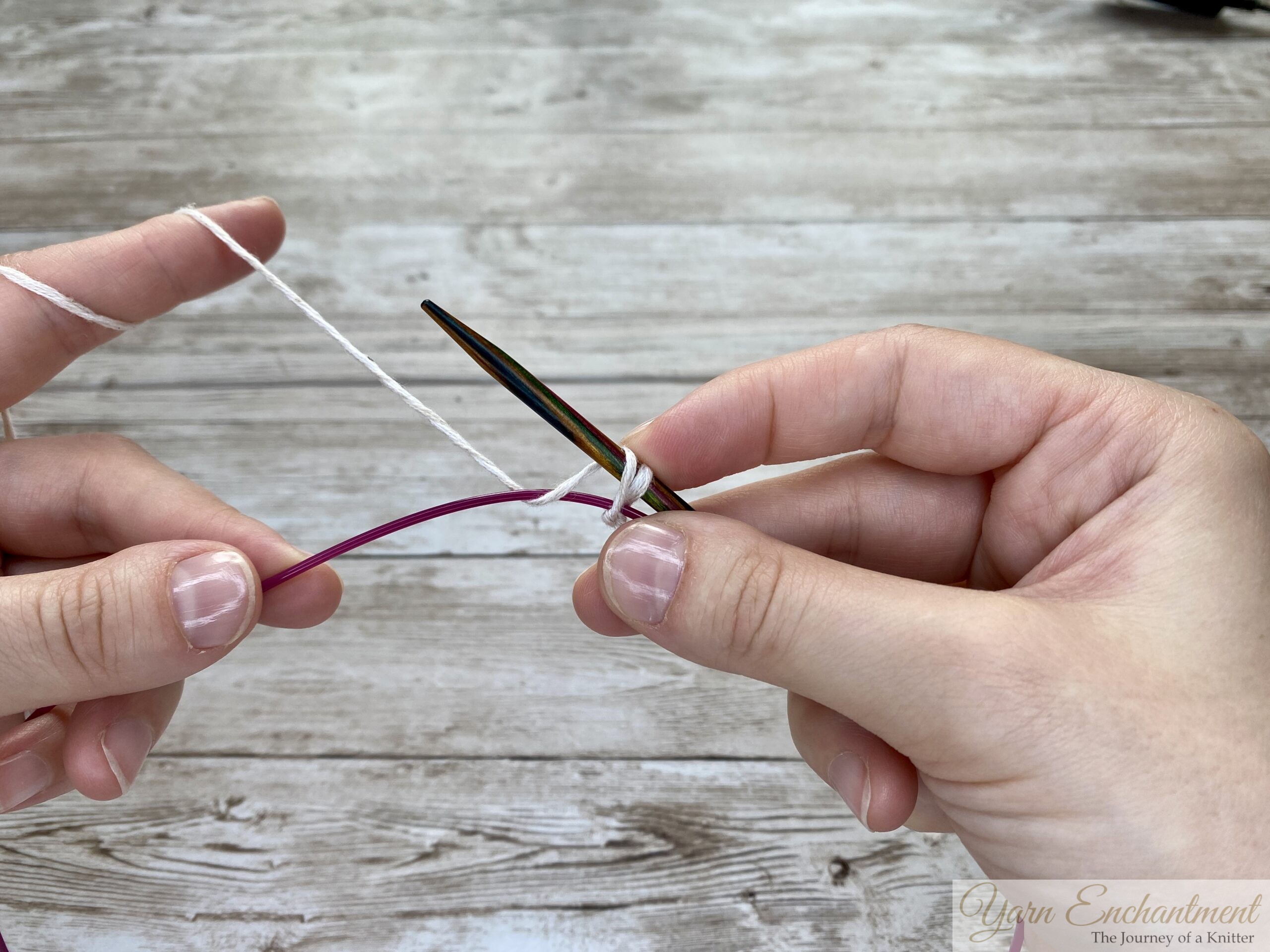Two hands demonstrating the provisional cast-on technique. The wooden knitting needle holds a few loops of white yarn, while the pink cable is positioned below to act as the spare holder for the cast-on stitches