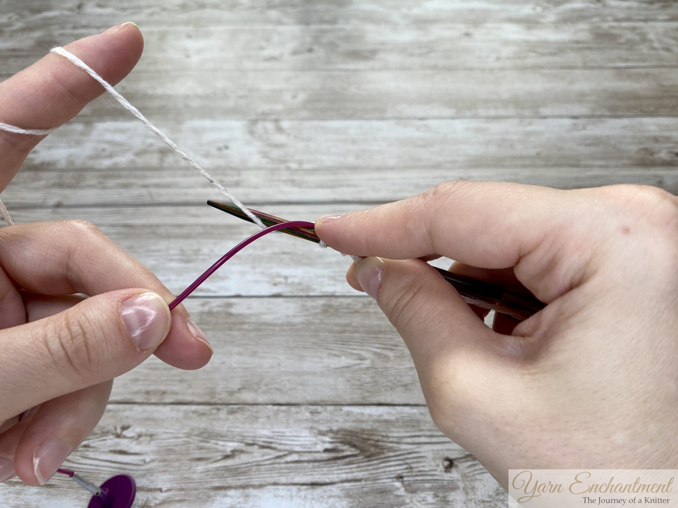 Two hands demonstrating the provisional cast-on technique. The wooden knitting needle holds a few loops of white yarn, while the pink cable is positioned below to act as the spare holder for the cast-on stitches