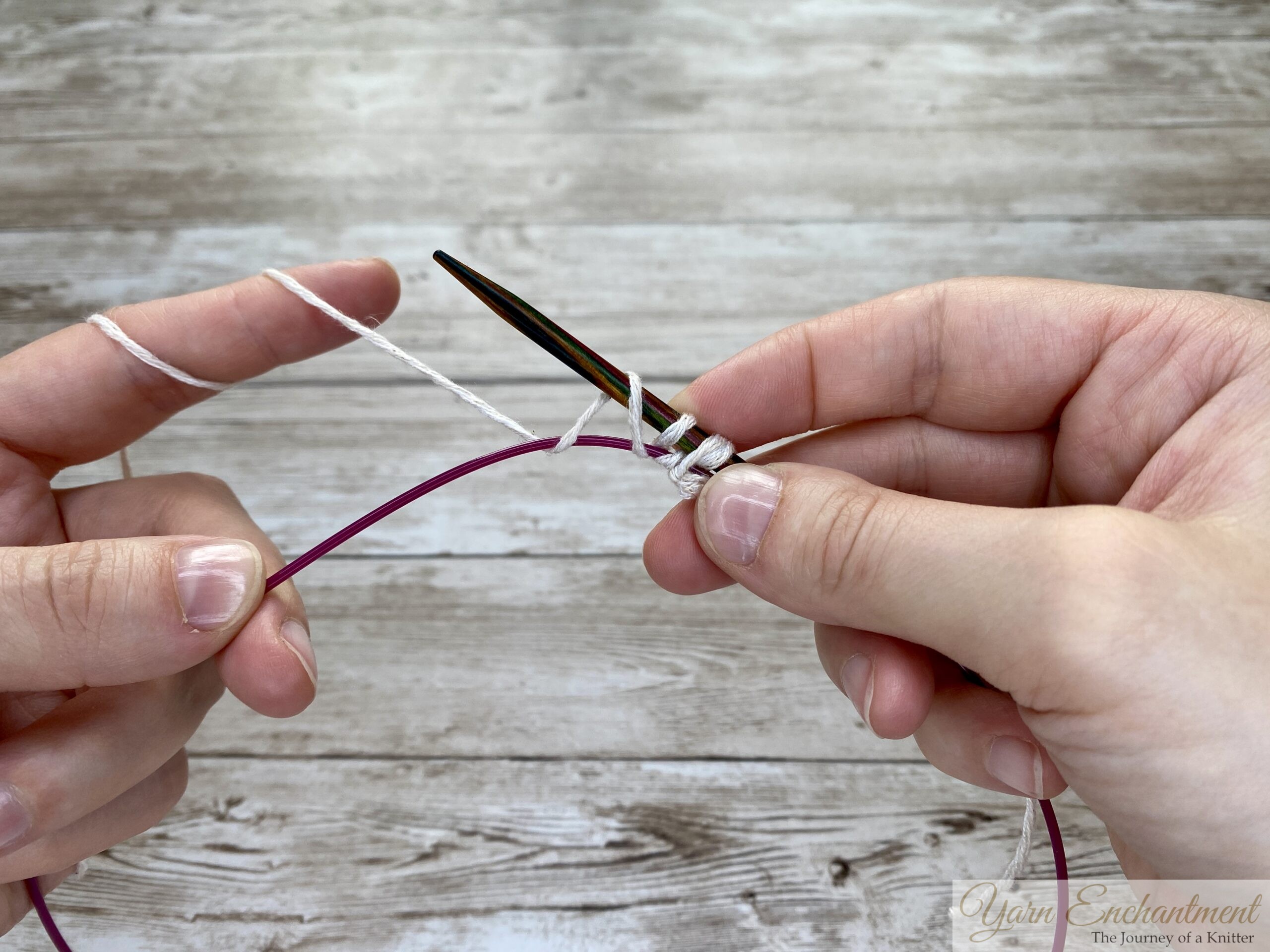 Two hands continuing the provisional cast-on process. The white yarn is being wrapped over the wooden needle, and the stitches are placed securely over the pink cable below