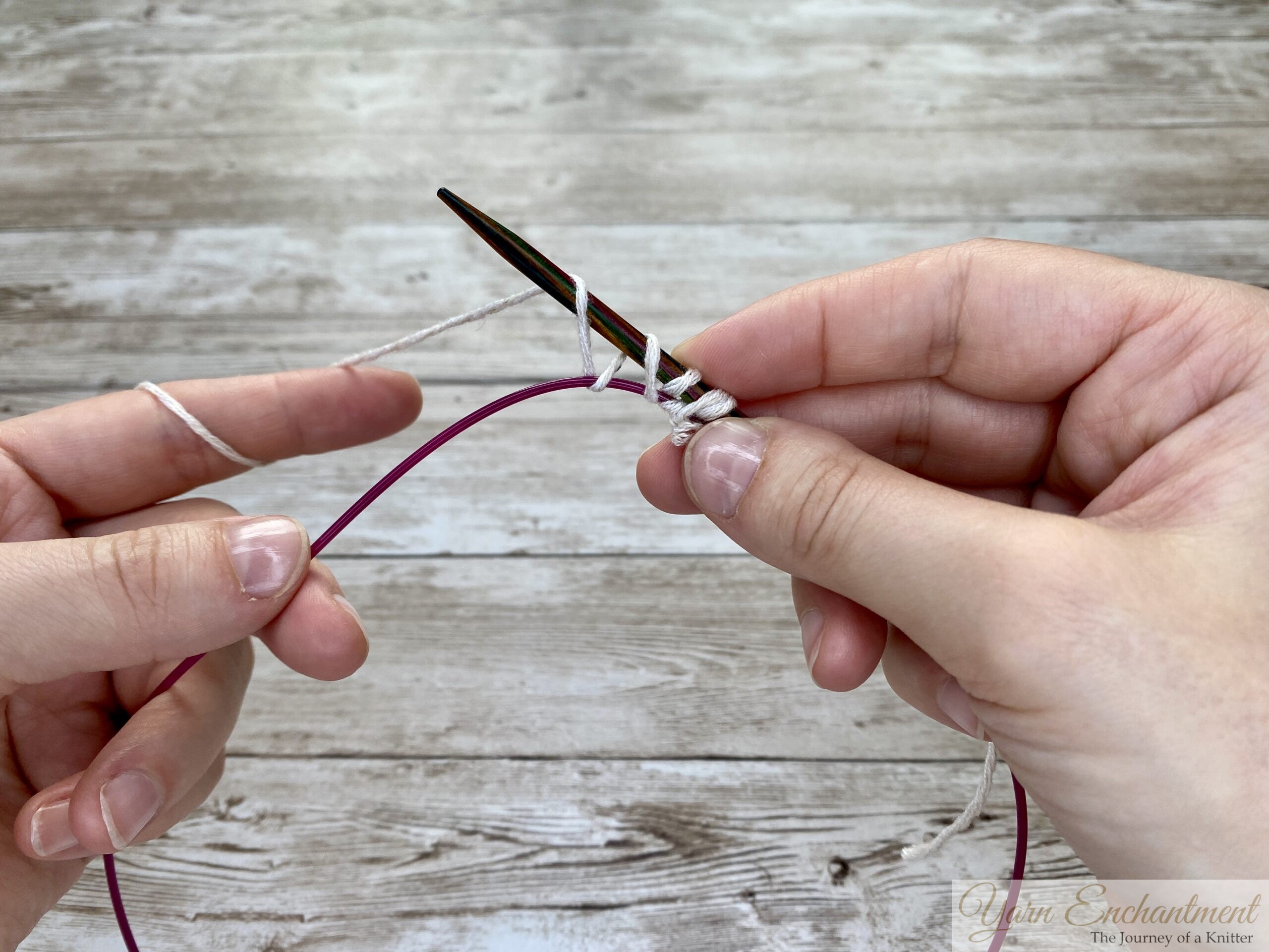 Two hands continuing the provisional cast-on process. The white yarn is being wrapped over the wooden needle, and the stitches are placed securely over the pink cable below