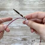 Progress on the provisional cast-on technique. Multiple loops of white yarn are visible on the wooden needle, with the pink cable below capturing and holding the provisional stitches securely