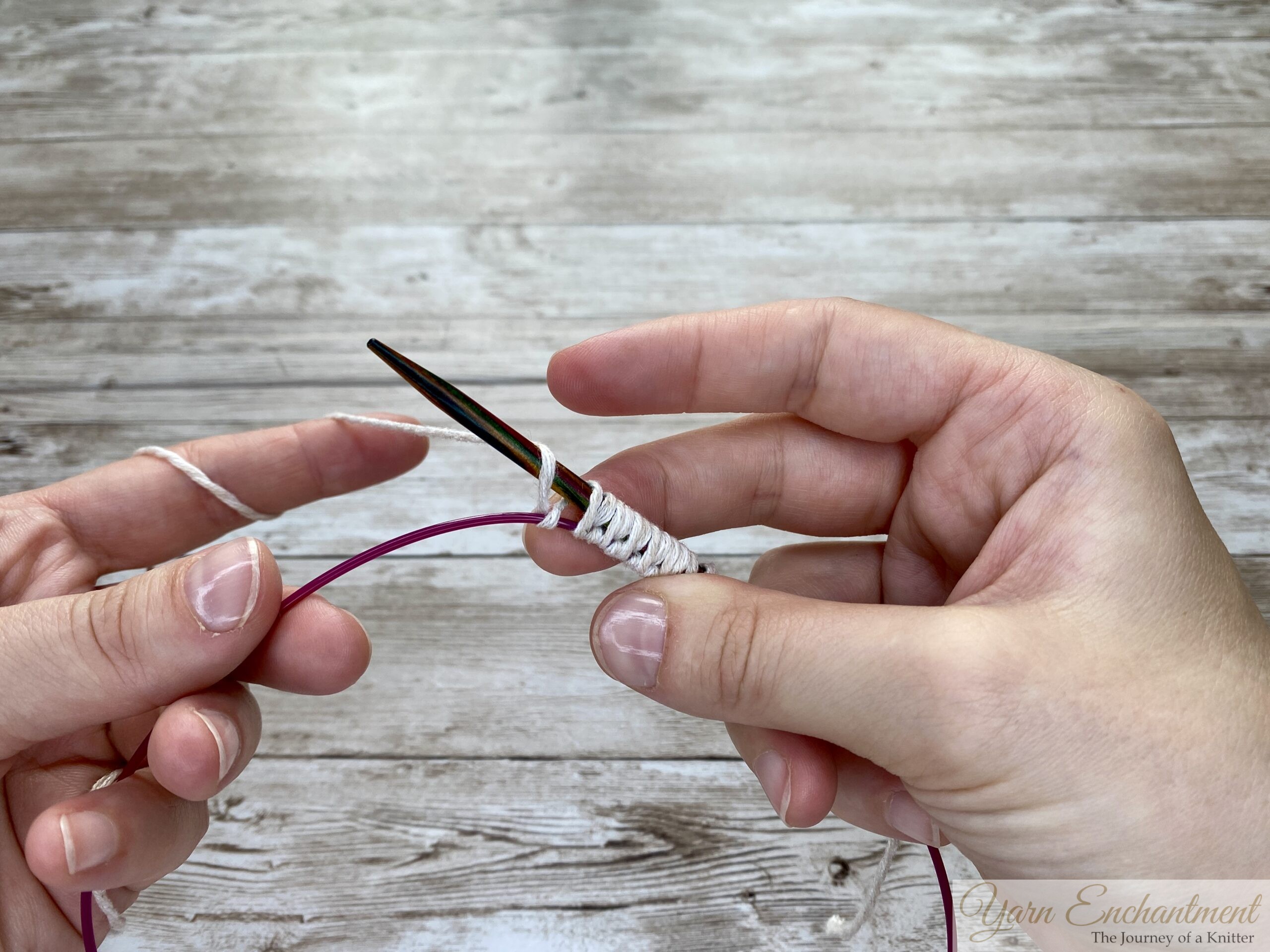 The provisional cast-on technique nearing completion. Several loops of white yarn are evenly wrapped around the wooden needle, with the pink cable acting as the spare holder