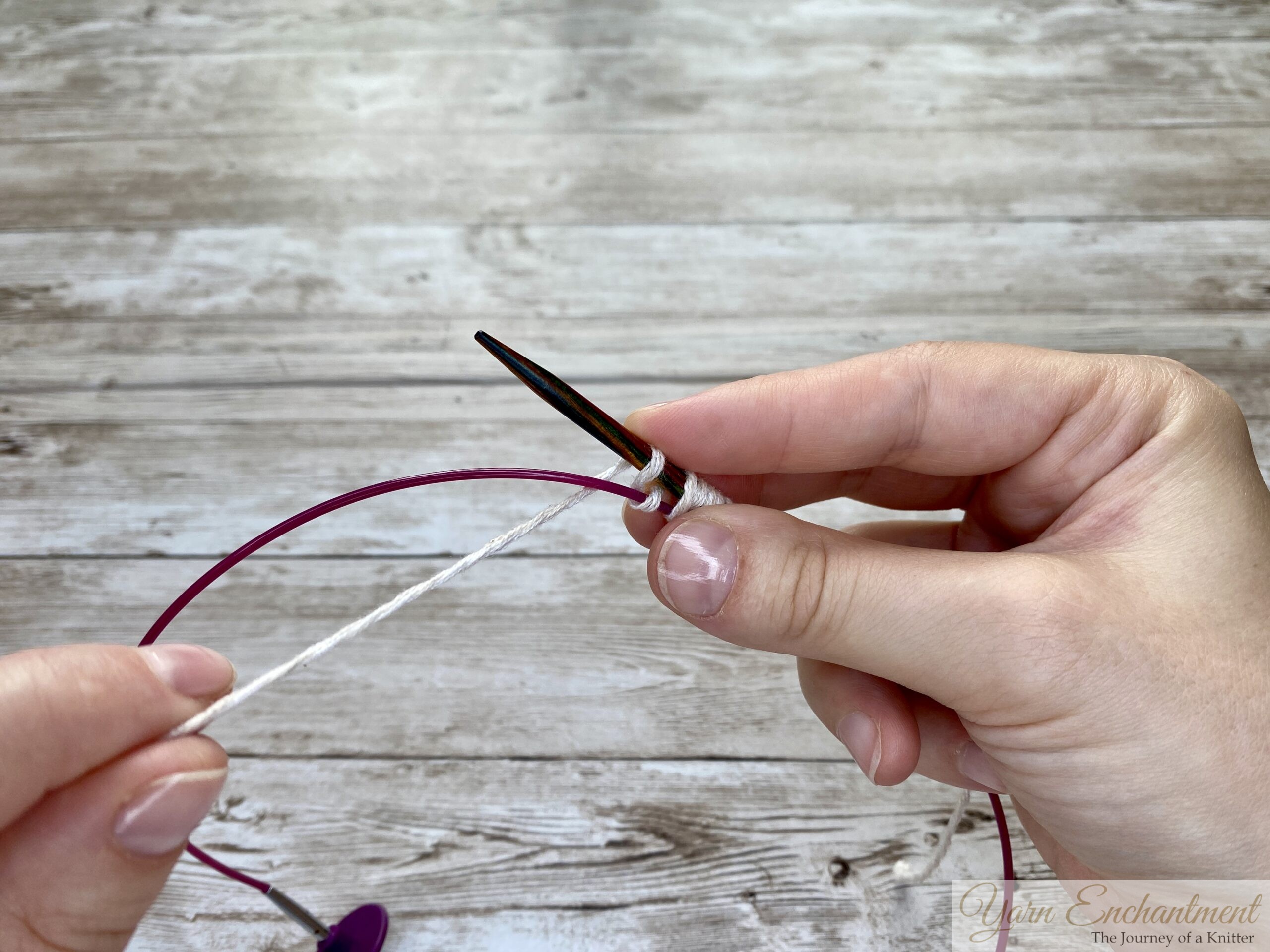 Two hands demonstrating the final stages of the provisional cast-on. The wooden needle holds the white yarn loops, and the pink cable below secures the provisional stitches.