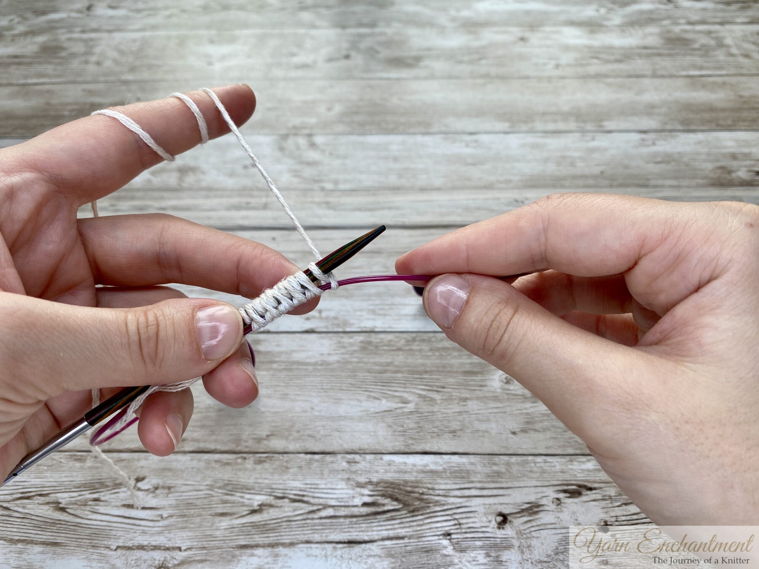 A close-up of the provisional cast-on technique. The wooden needle holds white yarn loops, with the left hand wrapping the yarn and the pink cable below capturing the stitches.