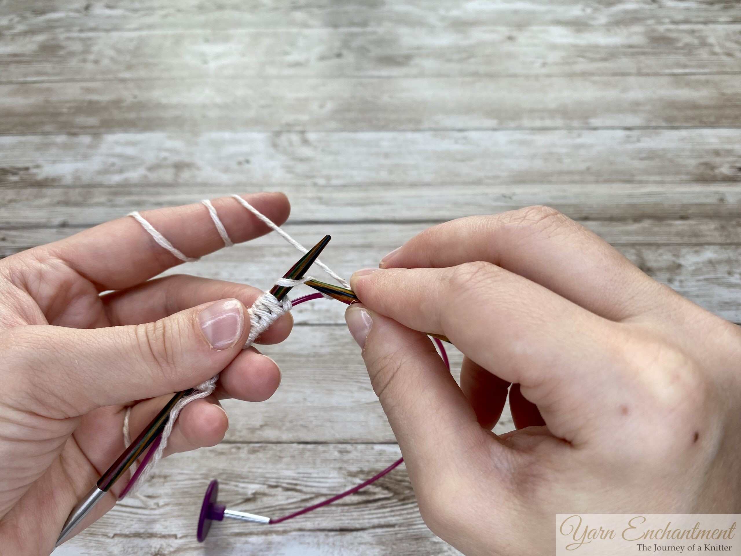 The wooden knitting needle holds the white yarn loops, and the second needle is introduced for further knitting, with the pink cable underneath still holding the provisional stitches.