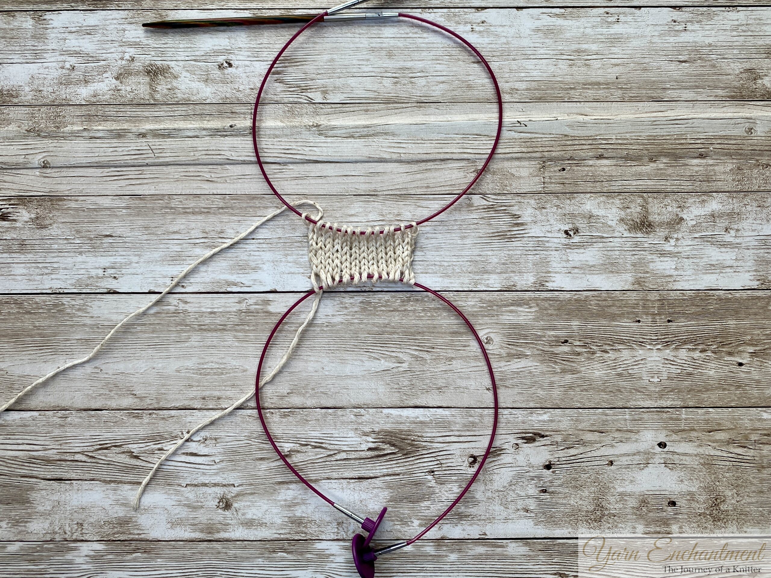 A completed swatch of white knitted fabric with stitches held securely on a pink cable and wooden needle. The setup shows the provisional cast-on in use, with the pink cable holding the initial stitches for future use.