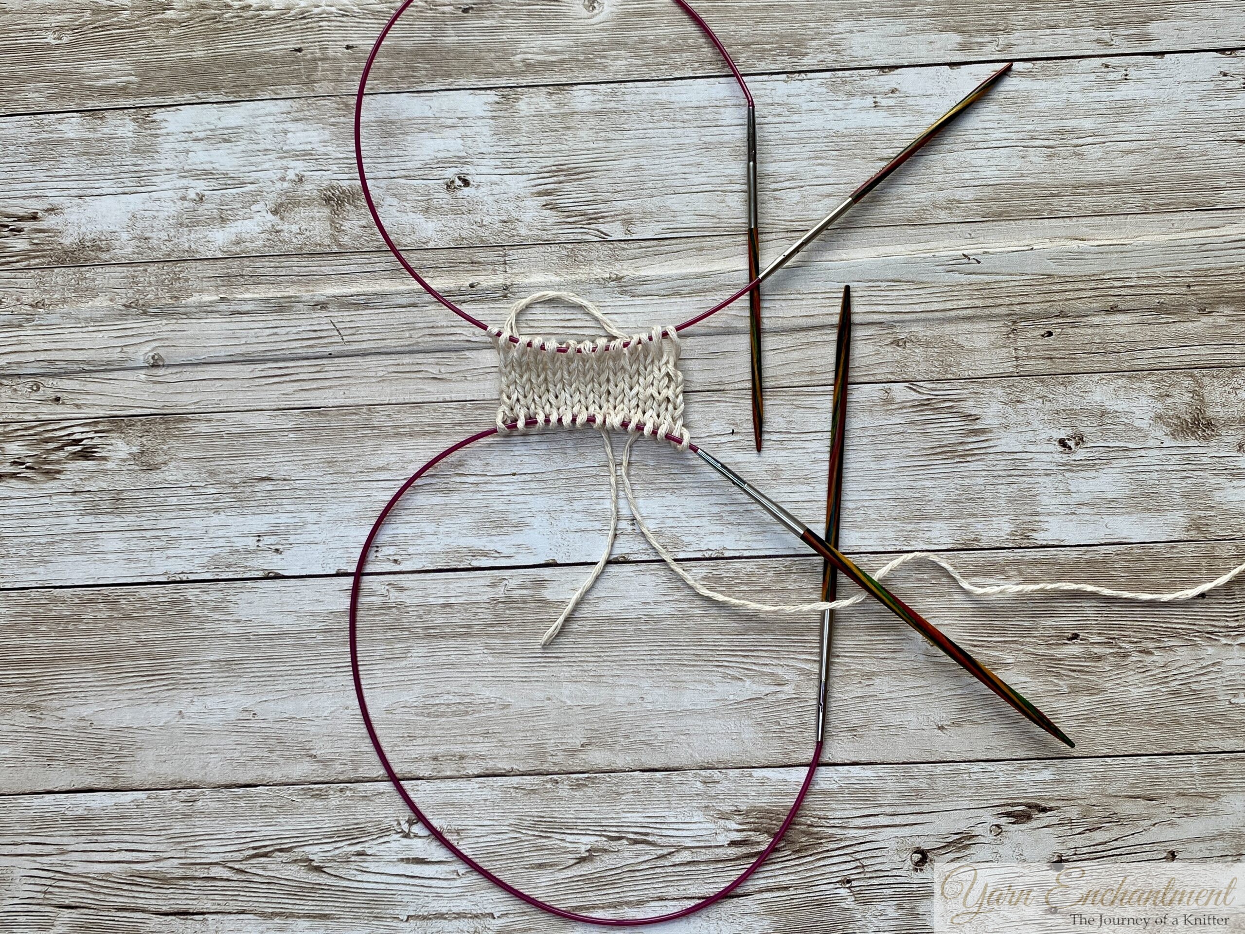 A top-down view of the completed provisional cast-on setup. The white knitted swatch is held between two pink cables and wooden needles, demonstrating readiness for future steps or adjustments in the project.