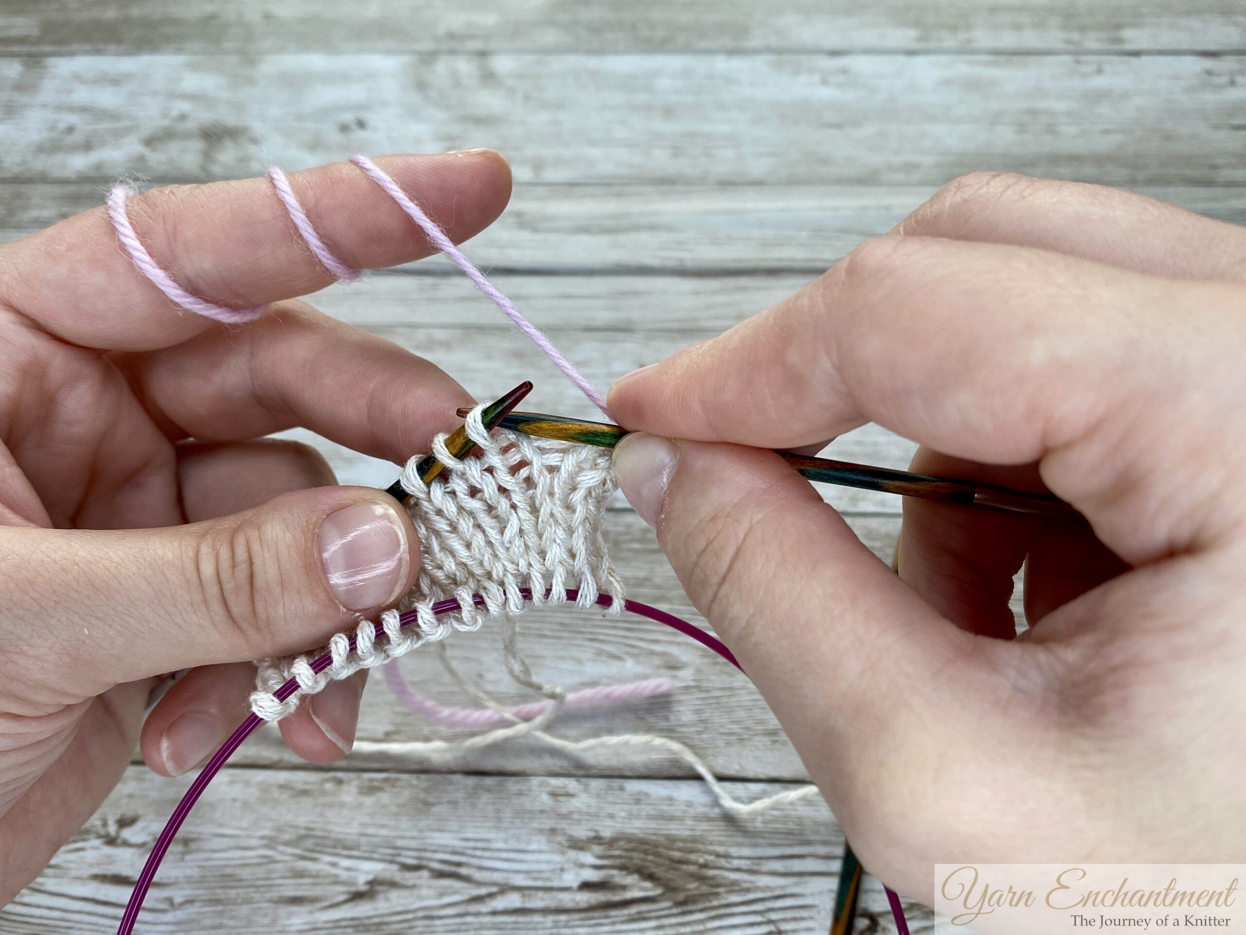 Two hands working on the knitted swatch. Pink yarn is being added to the white stitches held on the wooden needle, while the pink cable below secures the provisional cast-on stitches.