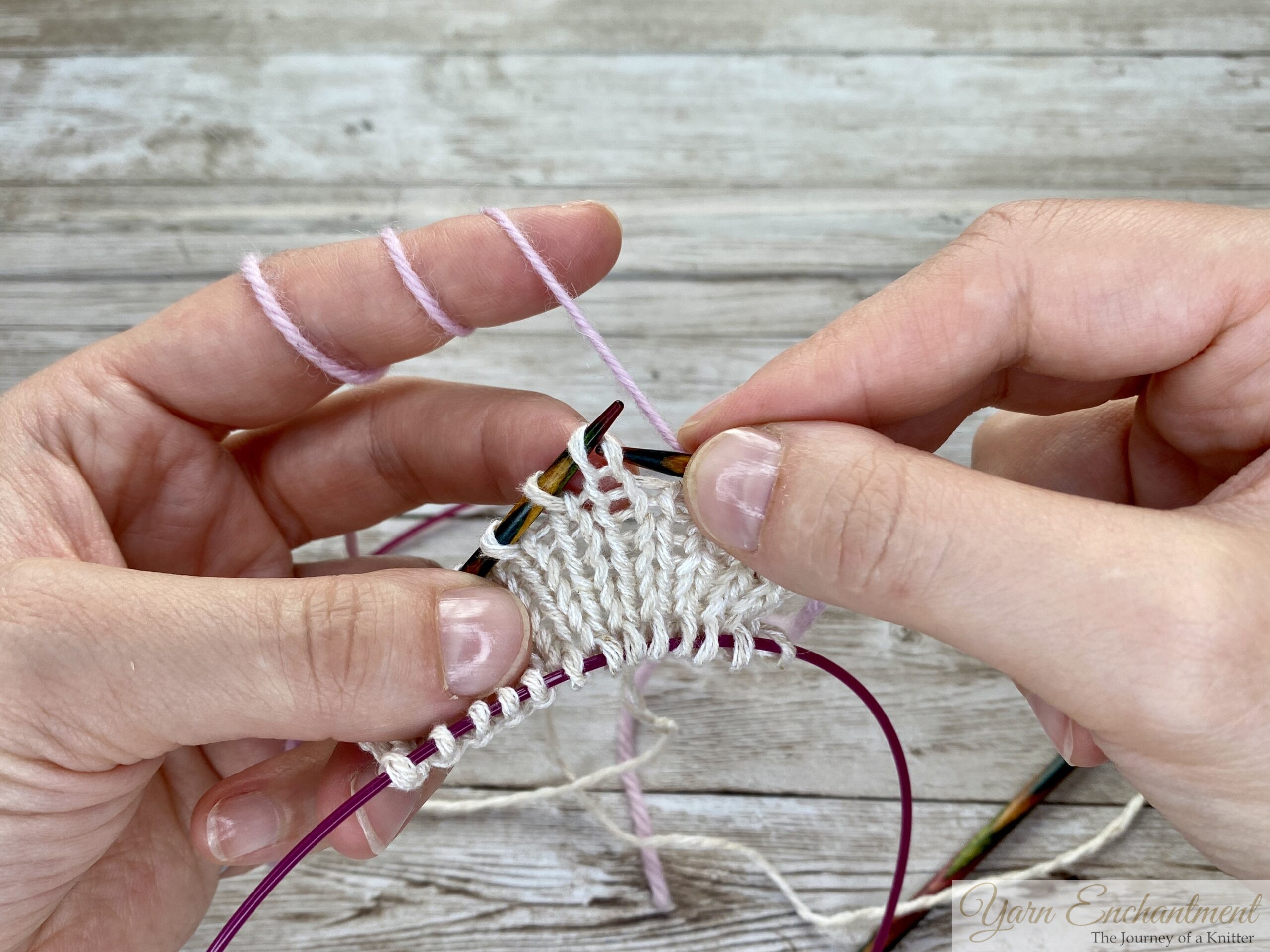 A close-up of the knitting process. Pink yarn is being incorporated into the white swatch, with the wooden needle actively working and the pink cable holding the bottom stitches.