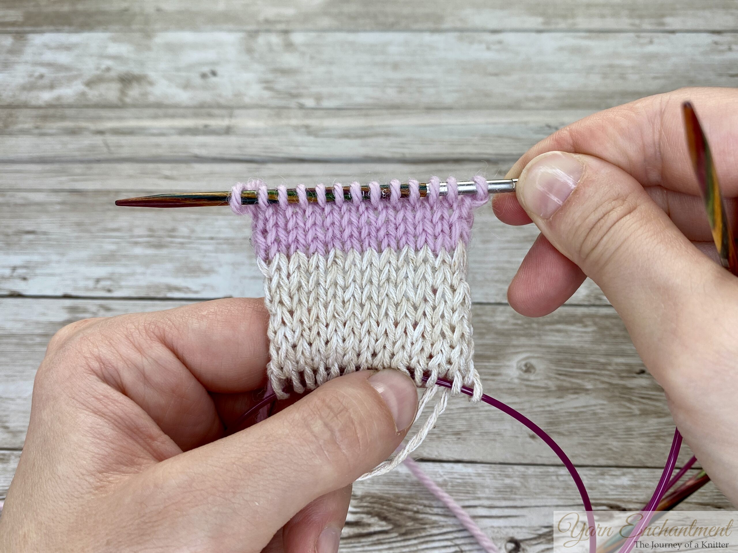 A completed swatch showing a transition between white and pink yarn. The stitches are neatly aligned on the wooden knitting needle, with the pink cable below holding the stitches
