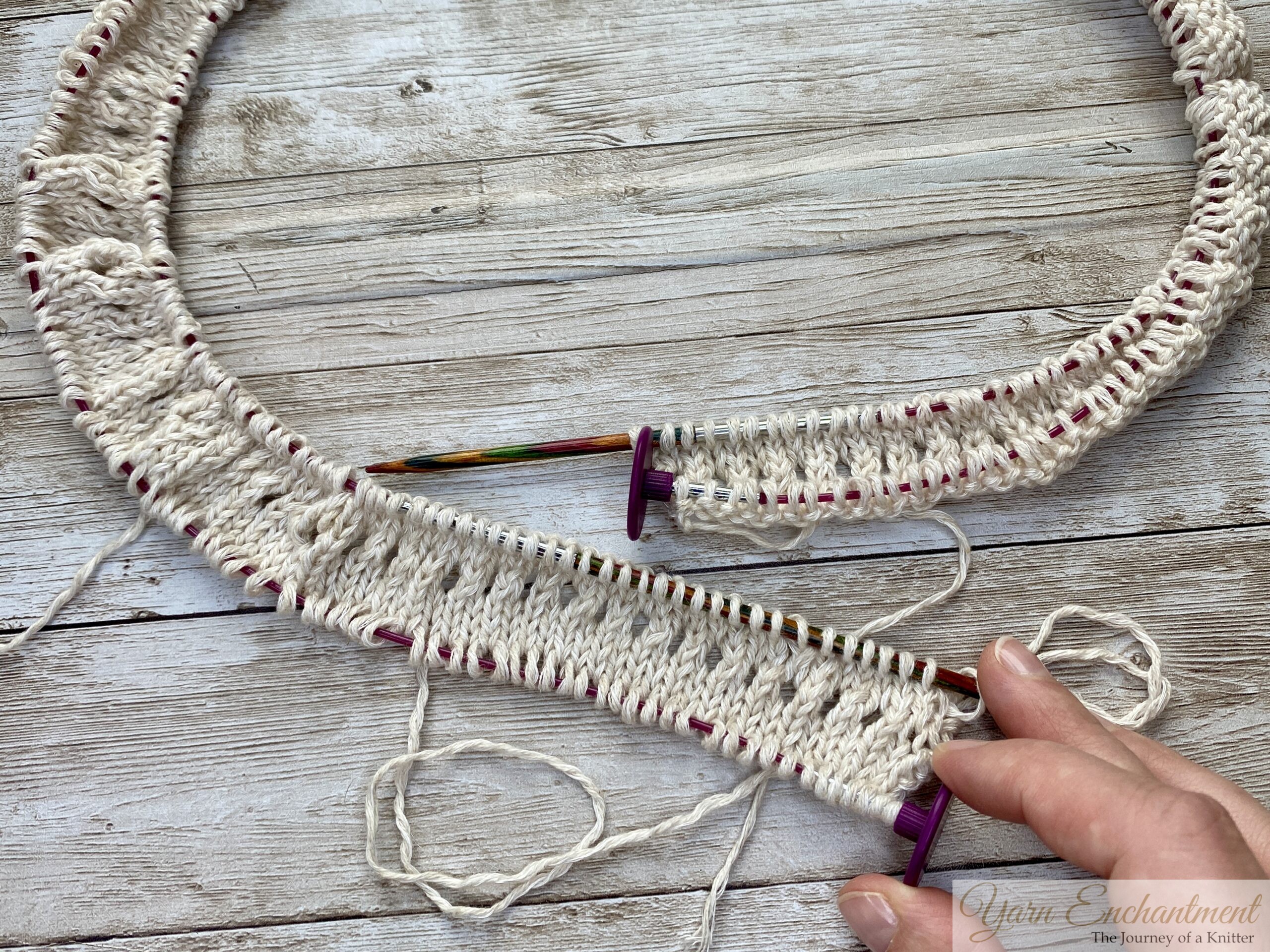 A larger knitted piece in progress using white yarn. The pink cable secures the provisional cast-on stitches and the wooden needle is actively working on the fabric