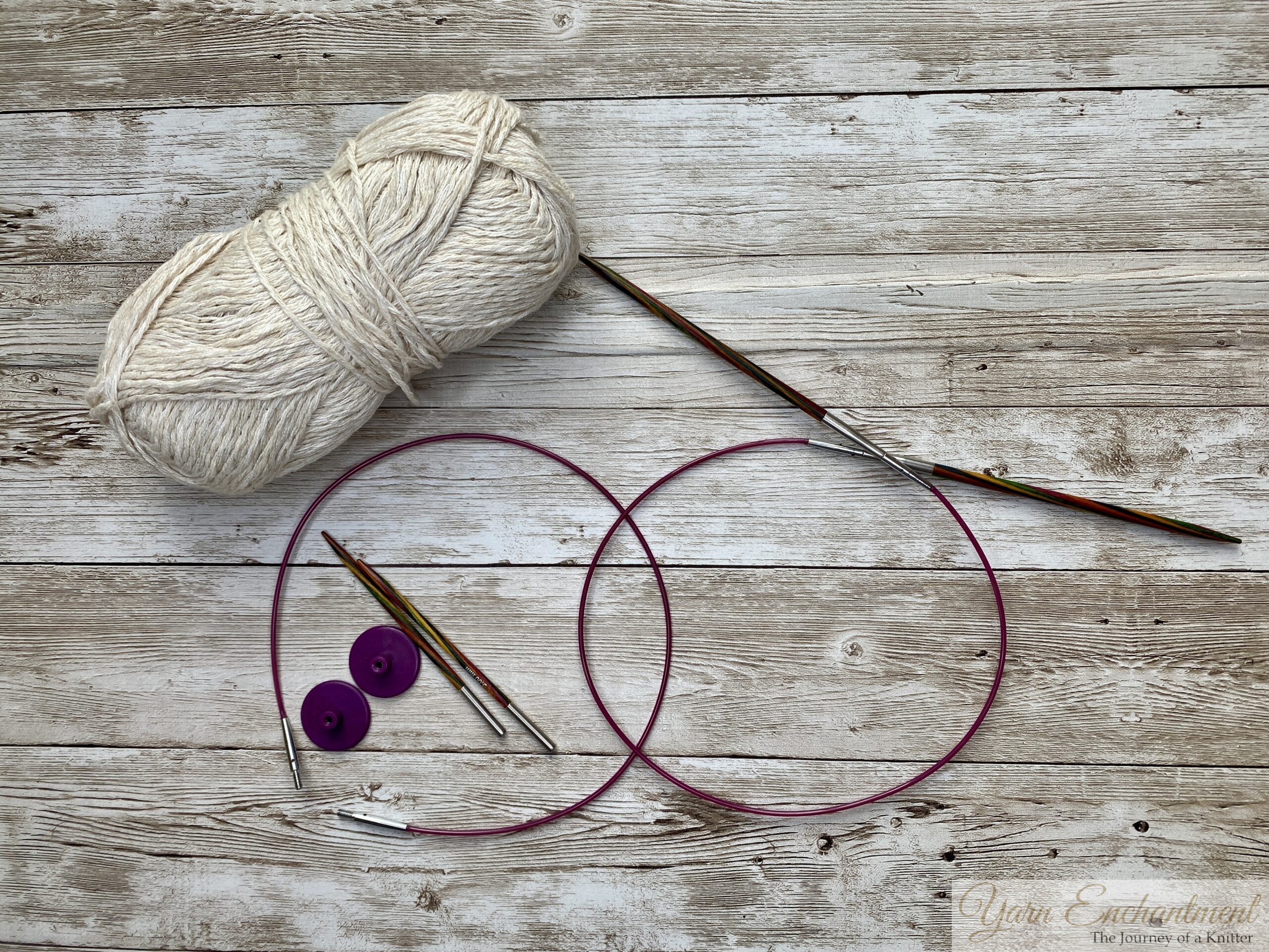 A flat-lay view of knitting essentials. A ball of white yarn, wooden needles, pink cables, and end caps are arranged on a wooden surface.