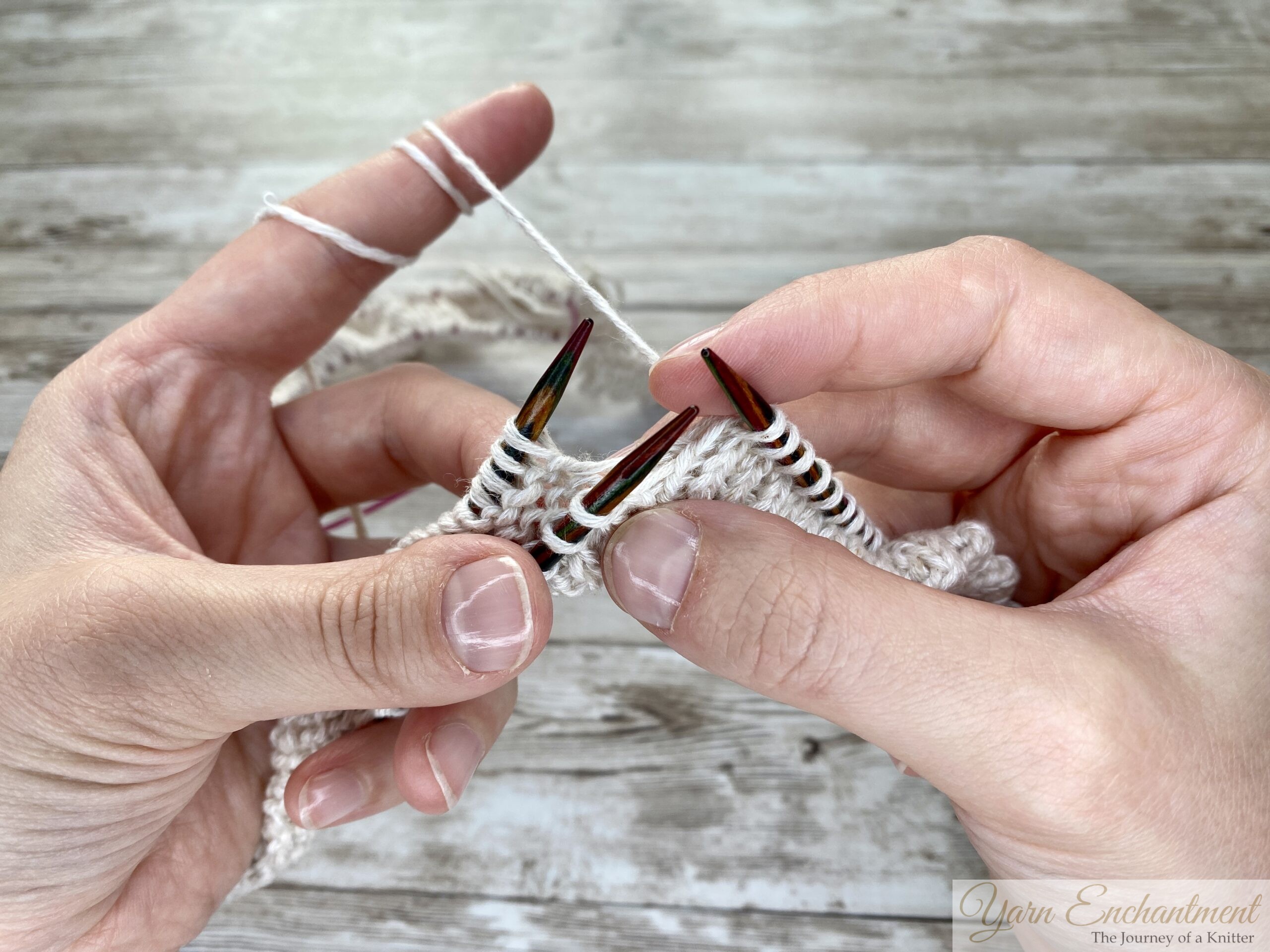 Active knitting in progress. The hands are working with white yarn on wooden needles, knitting two stitches together to create a folded edge