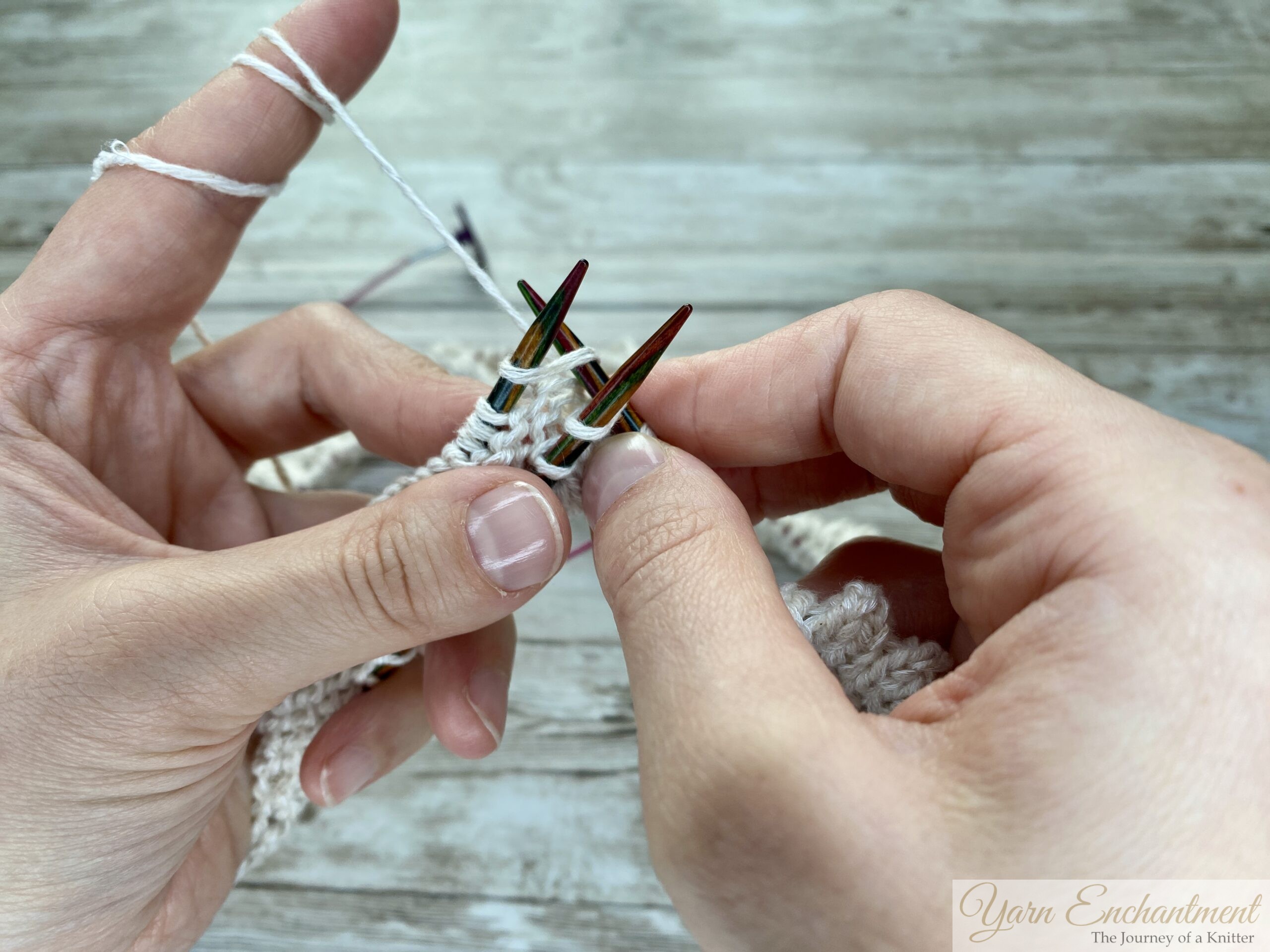 Active knitting in progress. The hands are working with white yarn on wooden needles, knitting two stitches together to create a folded edge