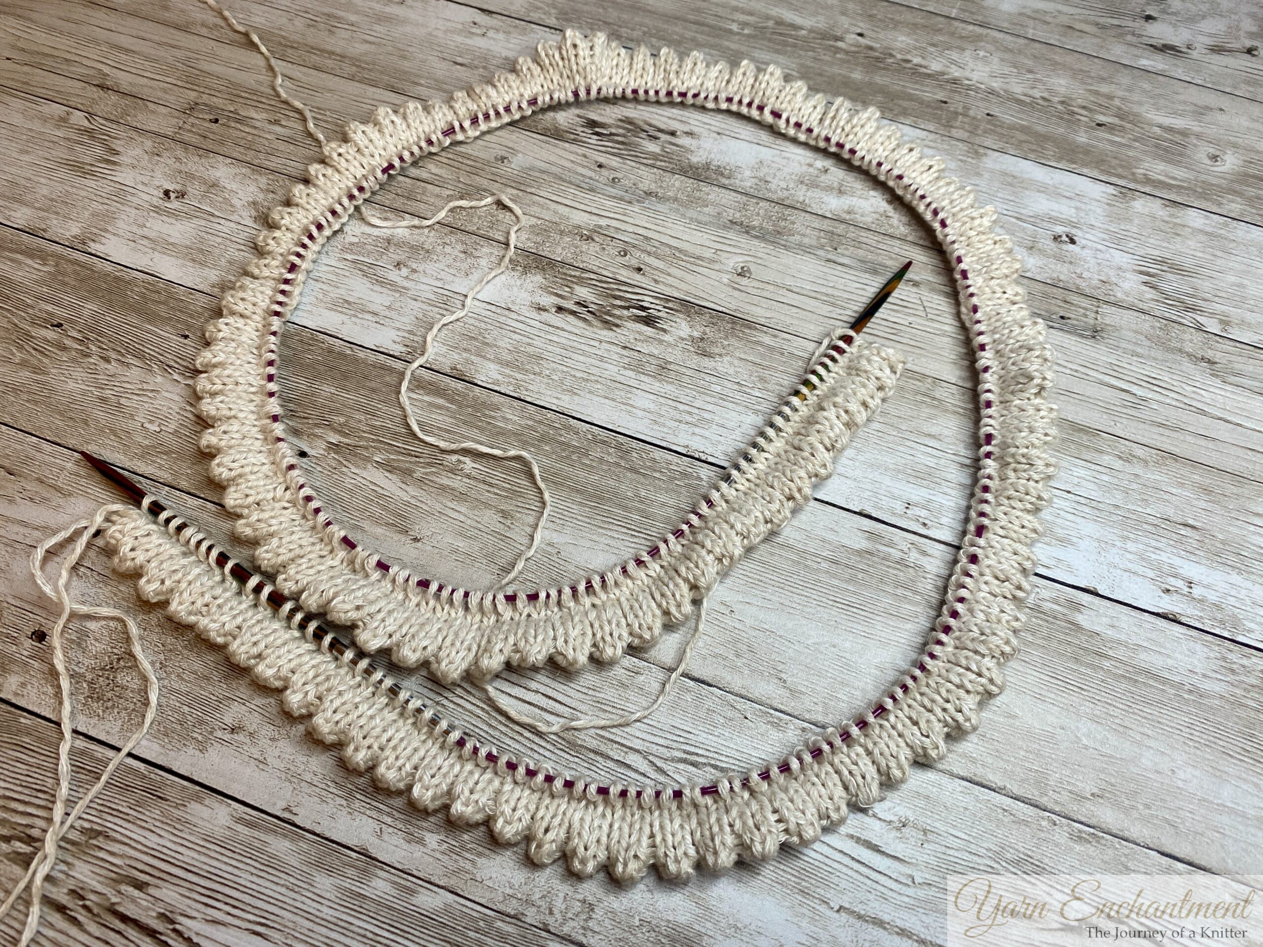Knitting project in progress using circular needles and cream-colored yarn with a double folded edge. The work is resting on a wooden surface with loose yarn visible.