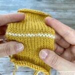Two hands holding a piece of yellow stockinette knitting with a white row of grafted stitches created using the Kitchener stitch. The seamless join is visible in the center, blending with the surrounding fabric.
