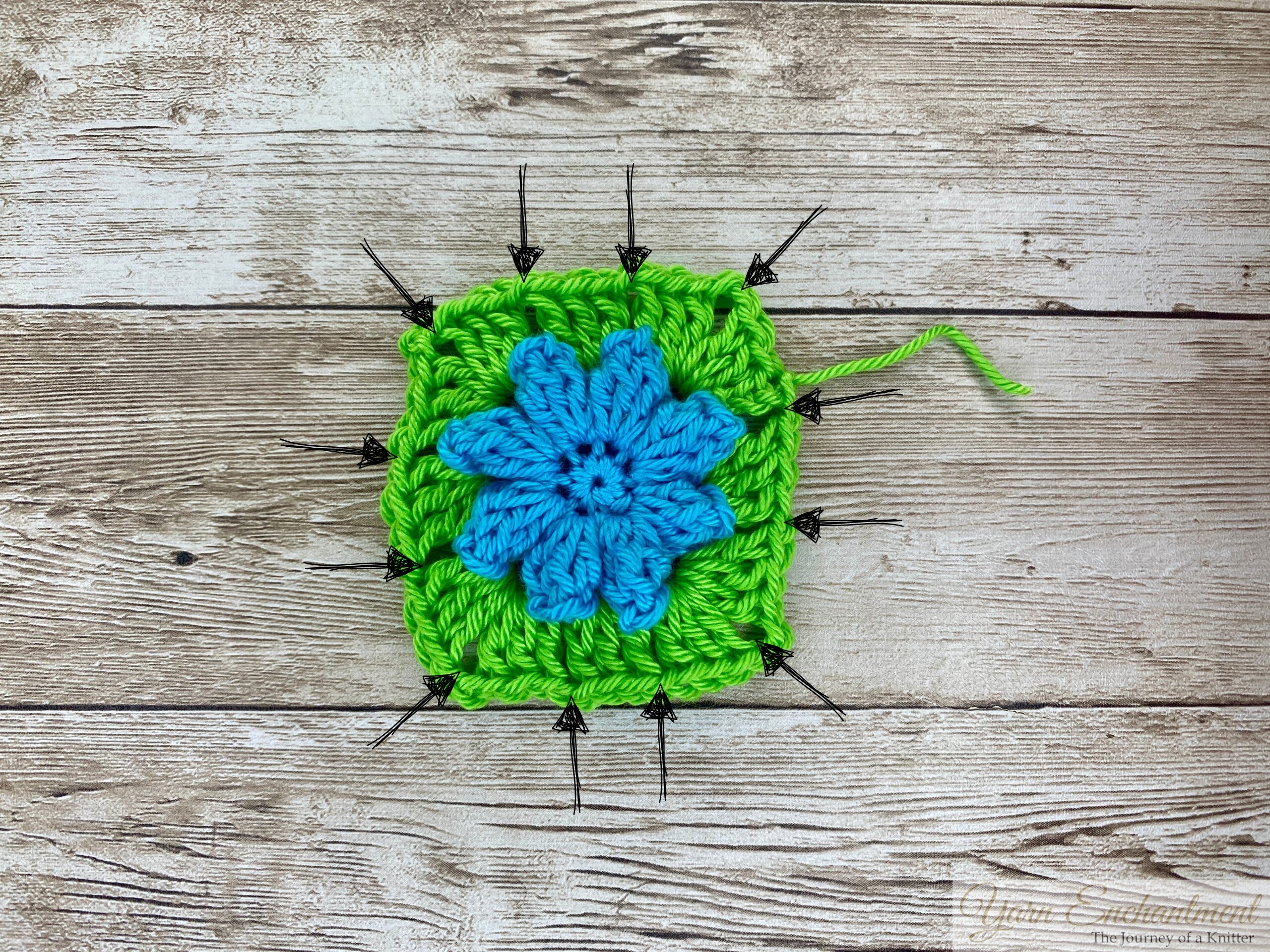 A completed crochet granny square featuring a bright blue flower centerpiece with green yarn surrounding it. The granny square is laid flat on a wooden surface.