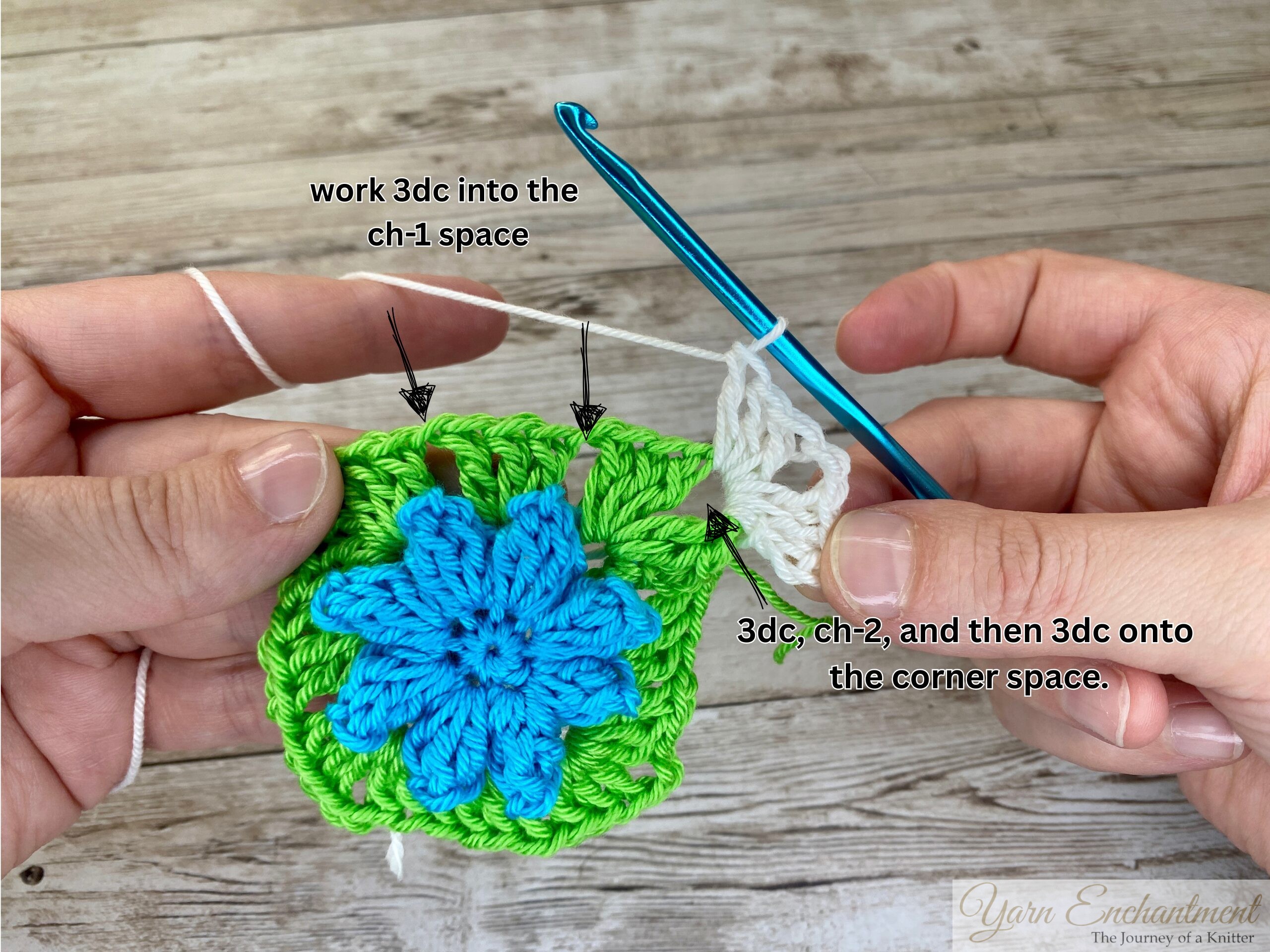 A crochet project in progress showing a blue flower centerpiece with green yarn forming a granny square. White yarn is being added to create a new border, with a blue crochet hook in use.