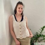 A handmade beige sleeveless knit top with wooden buttons and lace edging at the hem. The top is styled with neutral pants and a darker undershirt, complemented by a minimal indoor plant setup.