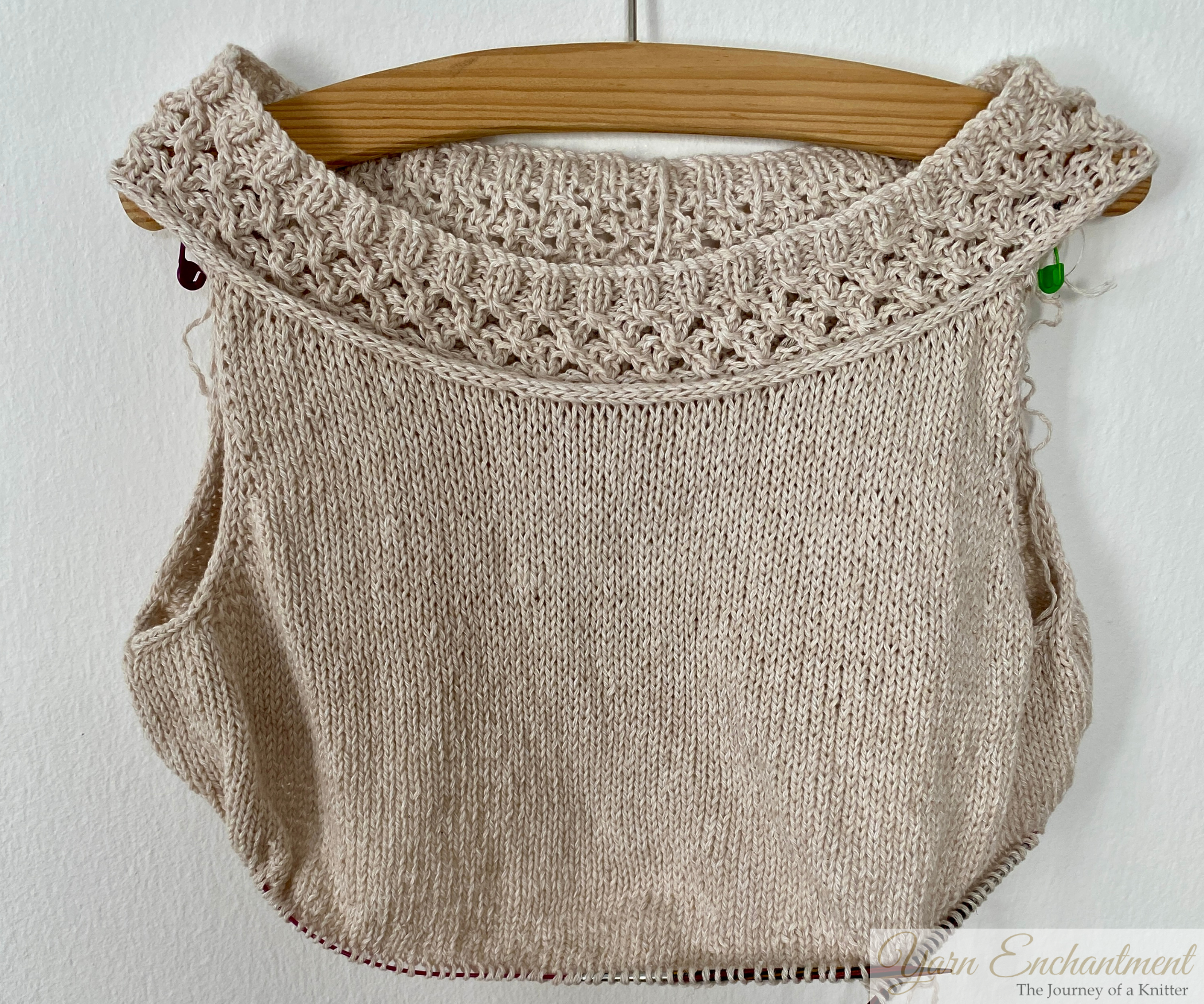 An unfinished beige knit garment with a lace yoke, displayed on a wooden hanger against a plain white wall. Circular knitting needles are still attached, indicating the work-in-progress state.