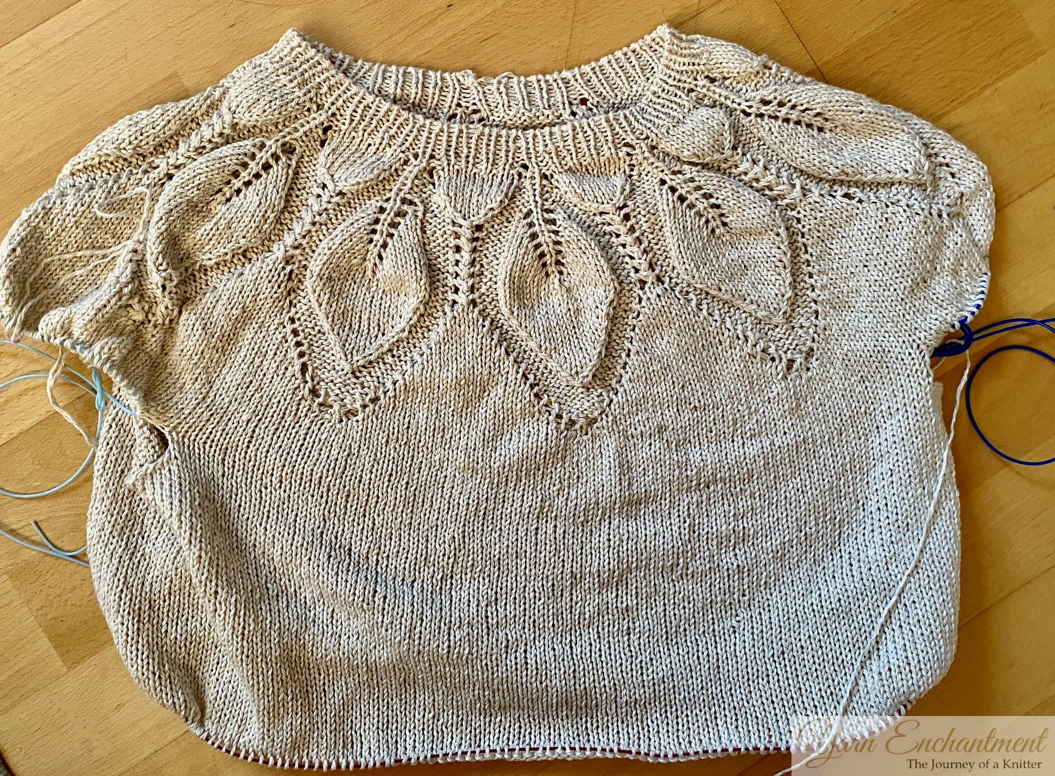 A knitted beige top with intricate leaf lace details on the yoke. The garment is laid flat on a wooden surface, showcasing its unfinished design with circular knitting needles still attached.