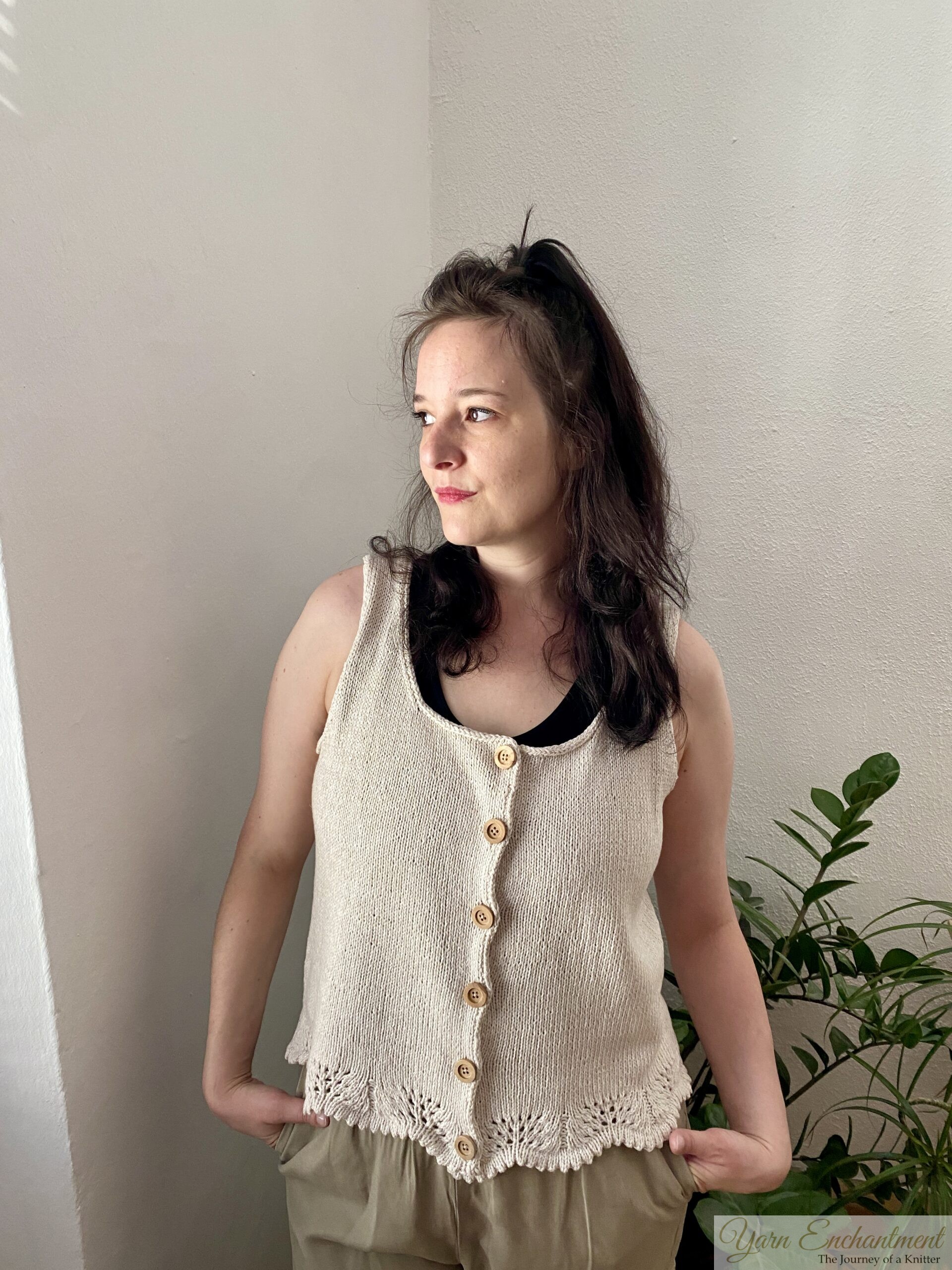 A handmade beige sleeveless knit top with wooden buttons and lace edging at the hem. The top is styled with neutral pants and a darker undershirt, complemented by a minimal indoor plant setup.