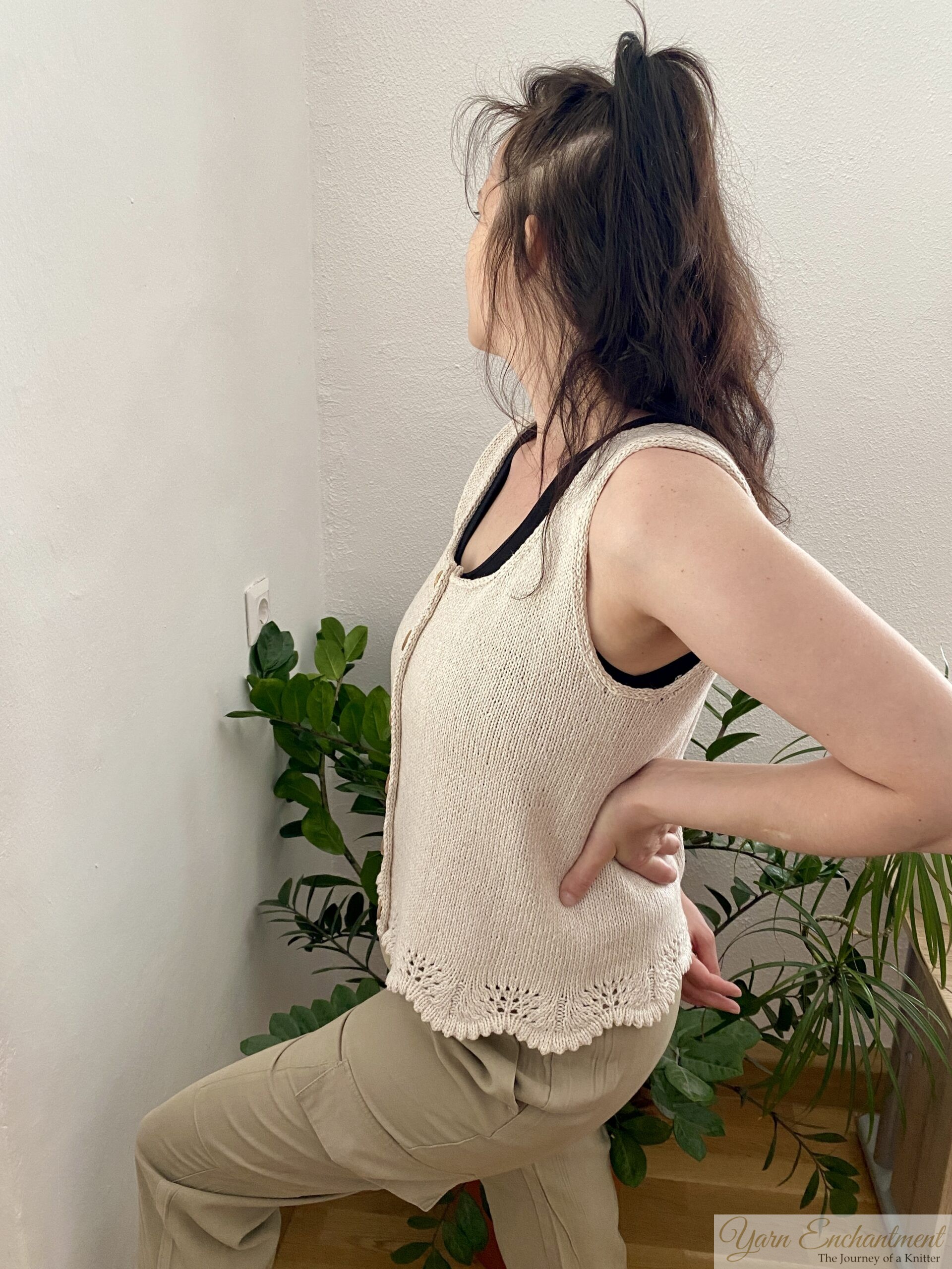 A person modeling a beige knitted sleeveless top with wooden buttons and lace detailing at the hem. The top is paired with neutral pants, and the background features green indoor plants and a minimalist wall.
