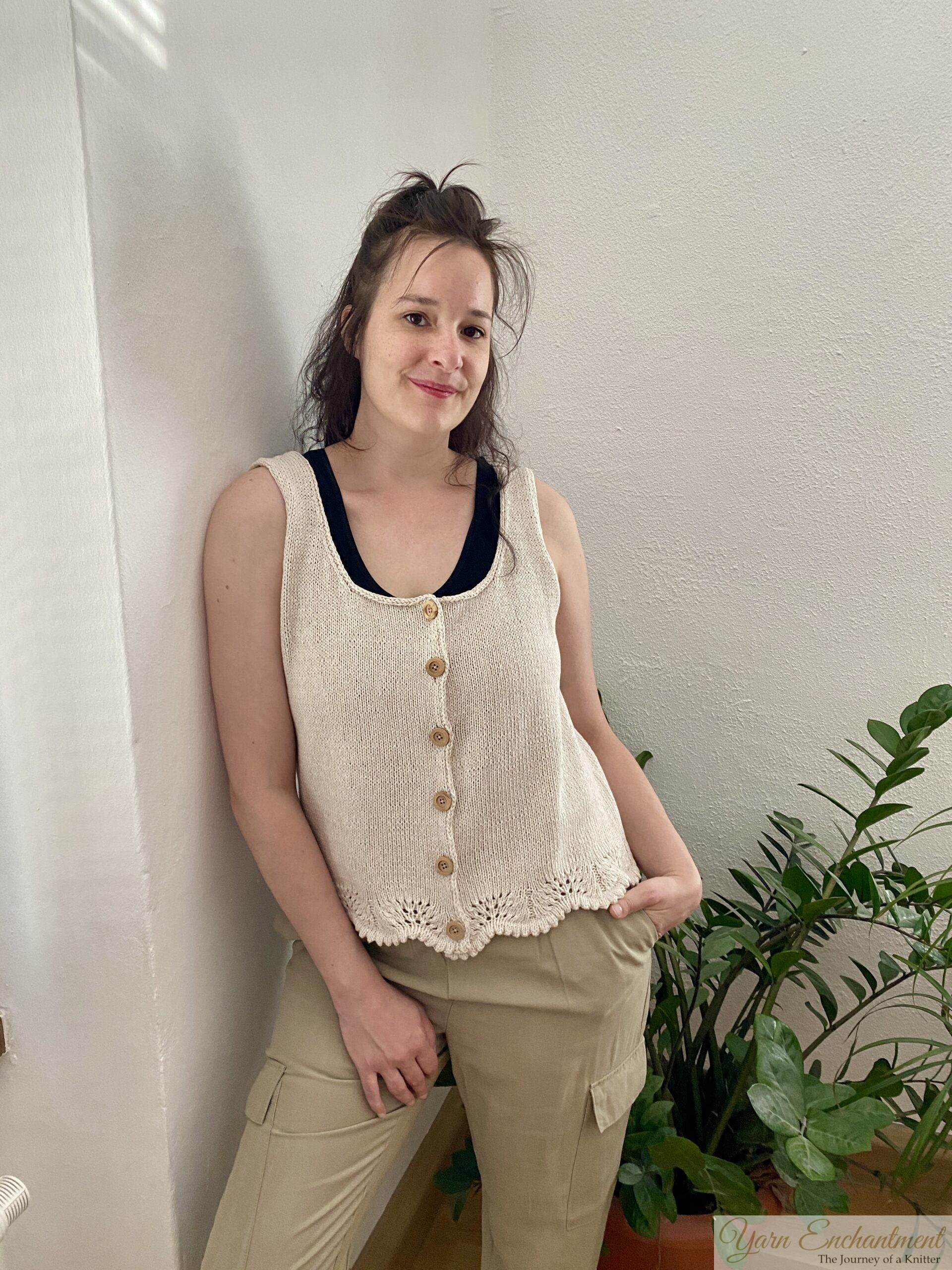 A handmade beige sleeveless knit top with wooden buttons and lace edging at the hem. The top is styled with neutral pants and a darker undershirt, complemented by a minimal indoor plant setup.