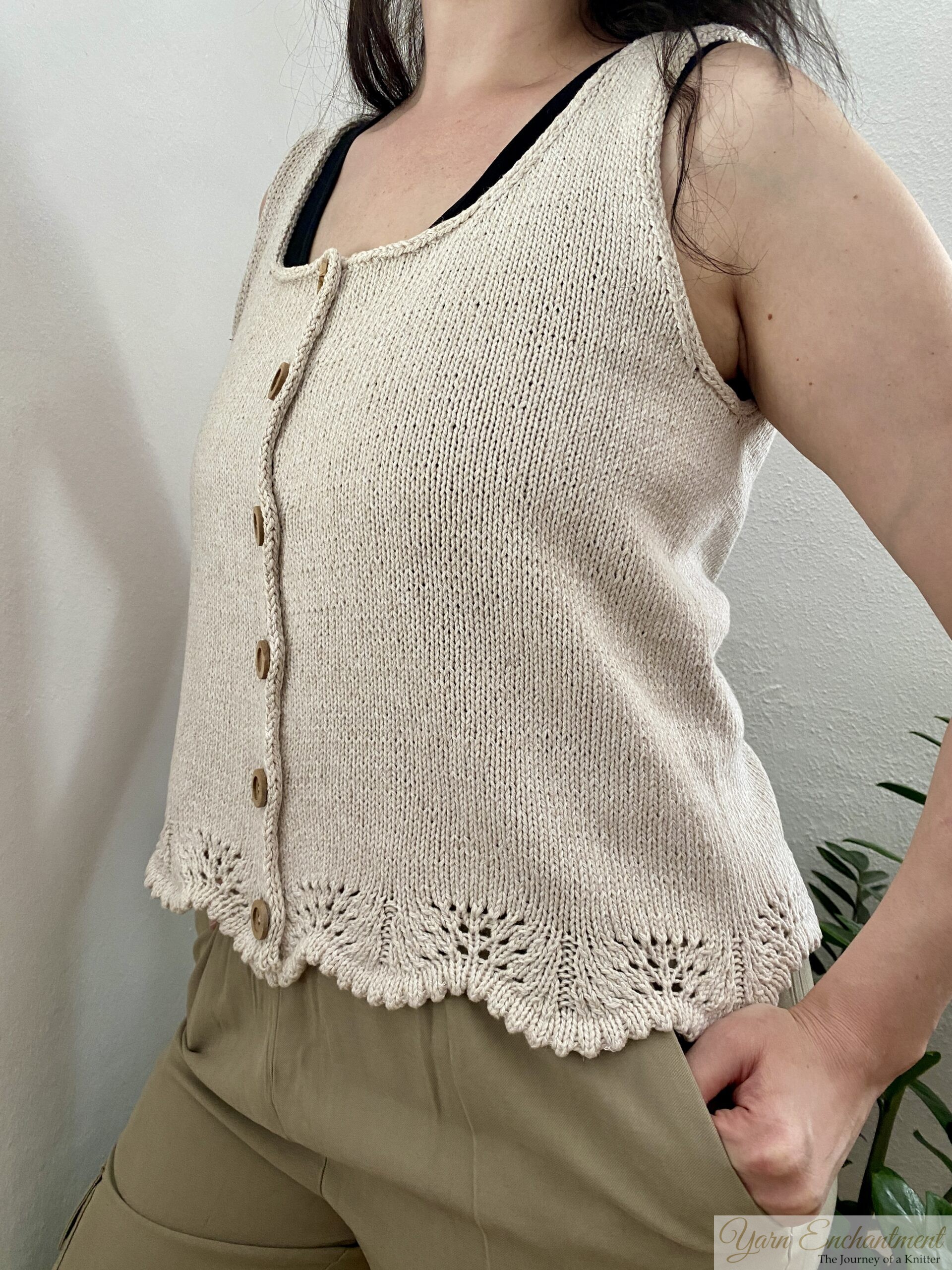 A handmade beige sleeveless knit top with wooden buttons and lace edging at the hem. The top is styled with neutral pants and a darker undershirt, complemented by a minimal indoor plant setup.