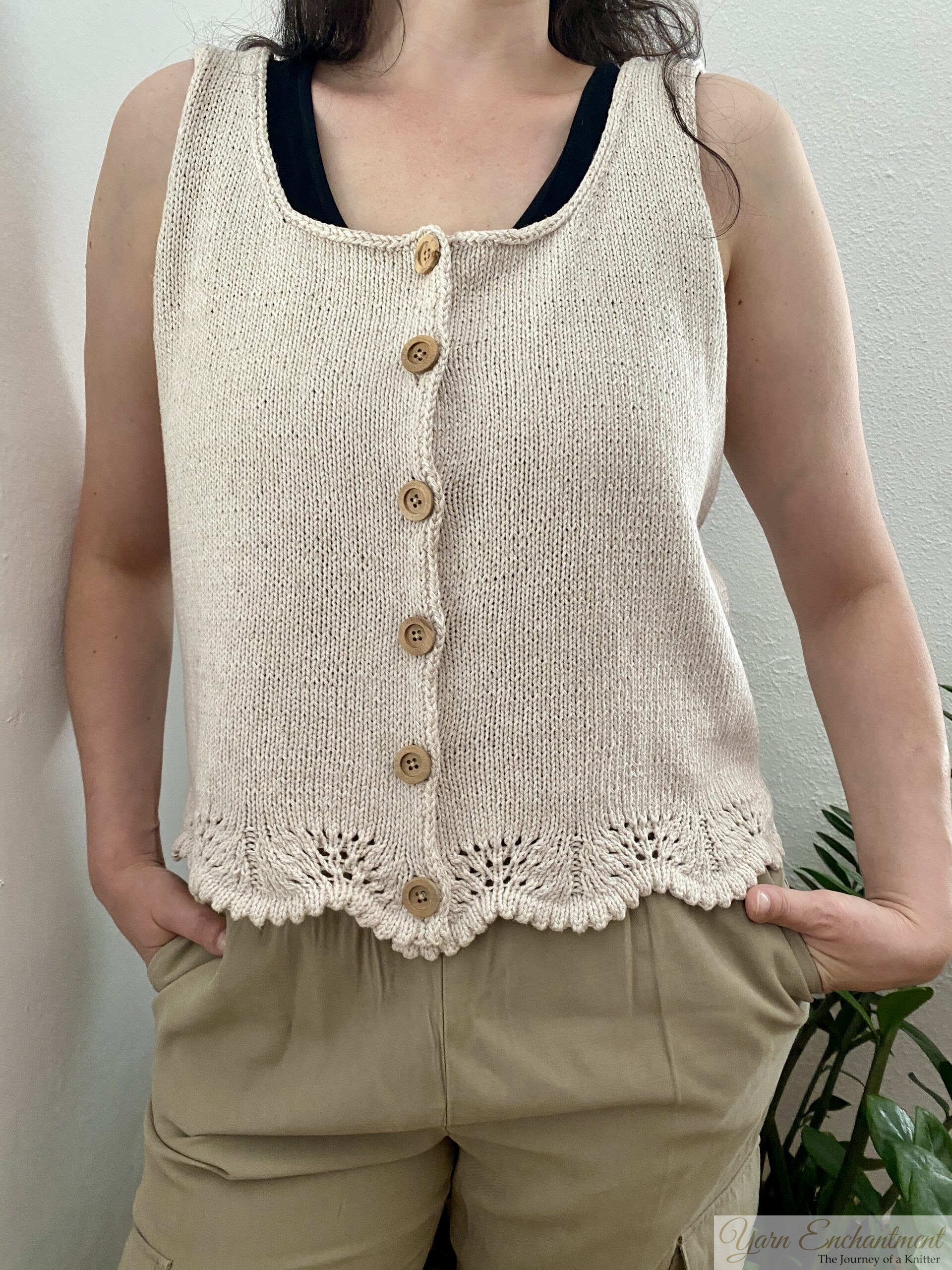 A handmade beige sleeveless knit top with wooden buttons and lace edging at the hem. The top is styled with neutral pants and a darker undershirt, complemented by a minimal indoor plant setup.
