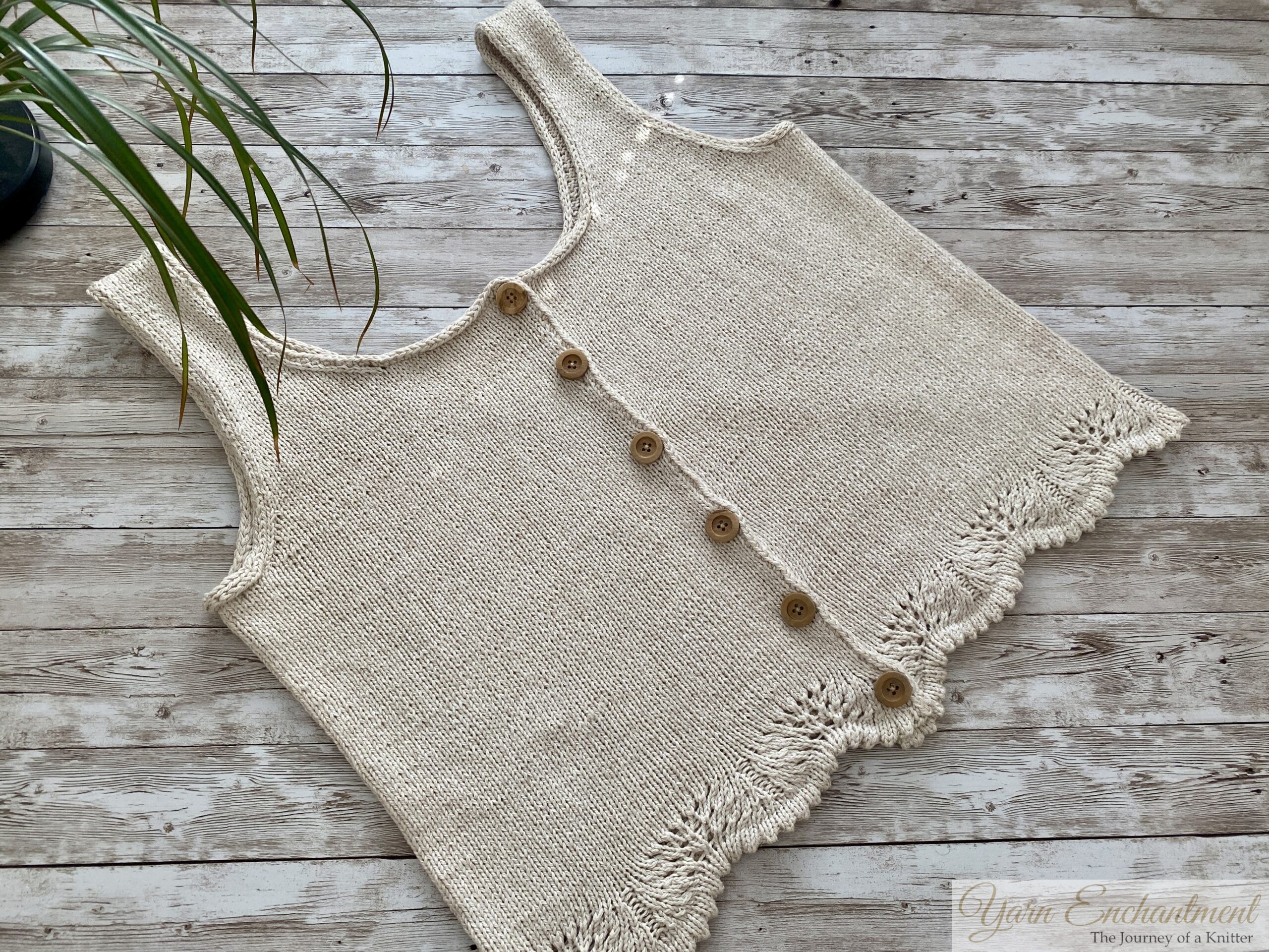 A beige sleeveless knit top with wooden buttons and lace detailing at the hem, laid flat on a wooden surface. The intricate lacework at the bottom is prominently displayed.
