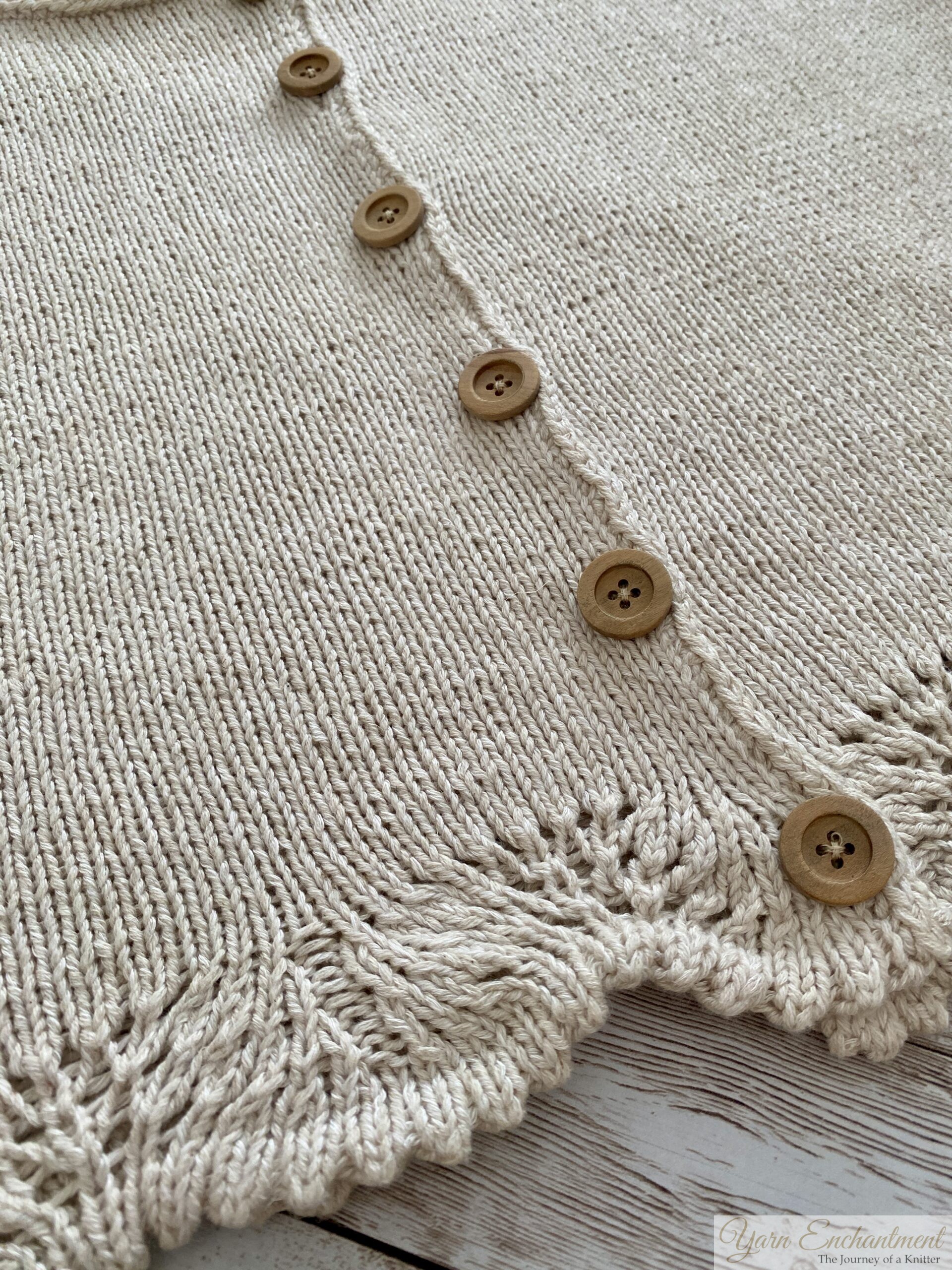 A close-up of a beige knitted top featuring wooden buttons running down the front and intricate lace detailing along the hem. The texture and stitchwork of the fabric are highlighted.