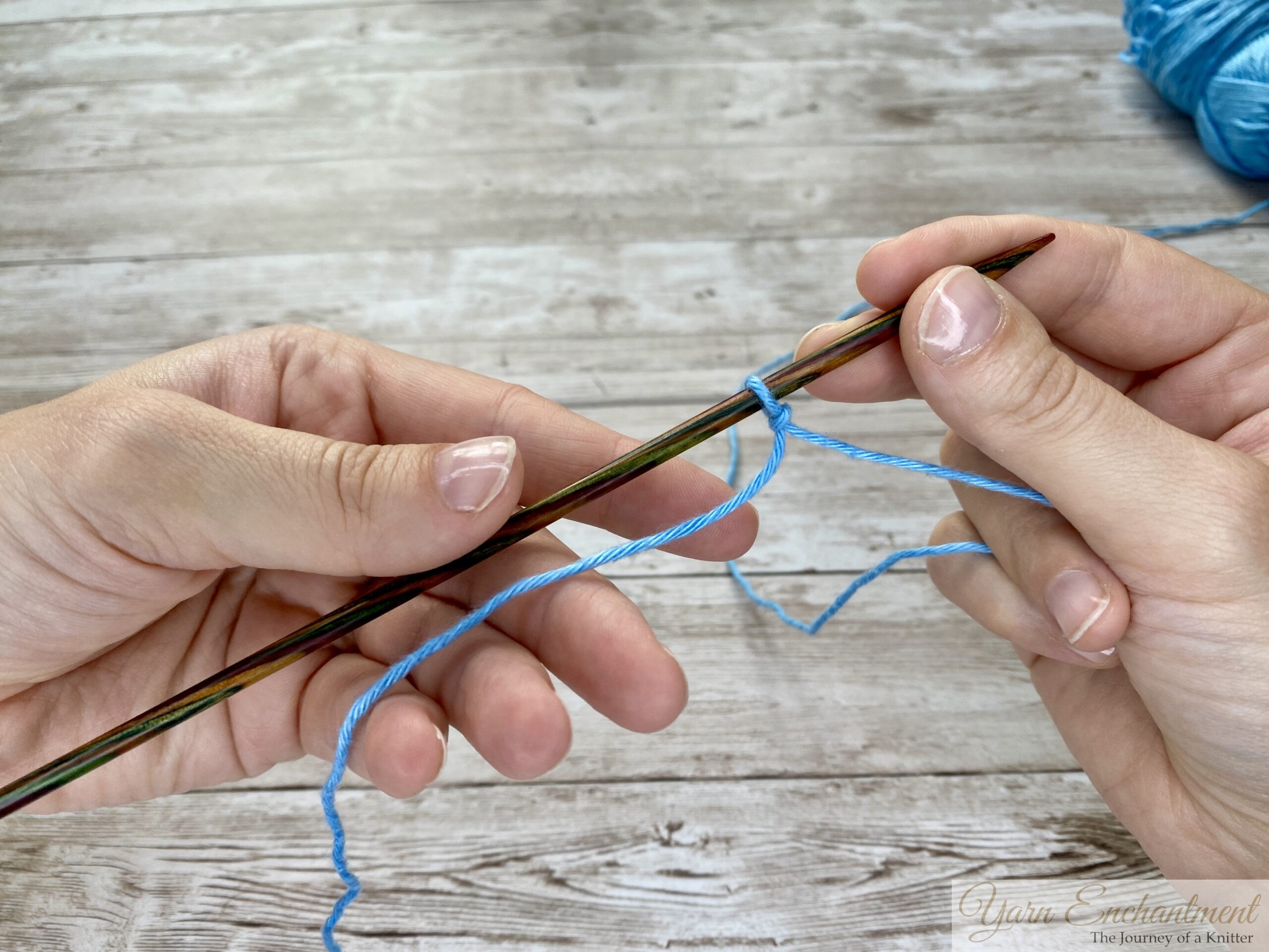 Two hands holding a knitting needle with blue yarn looped, ready for casting on