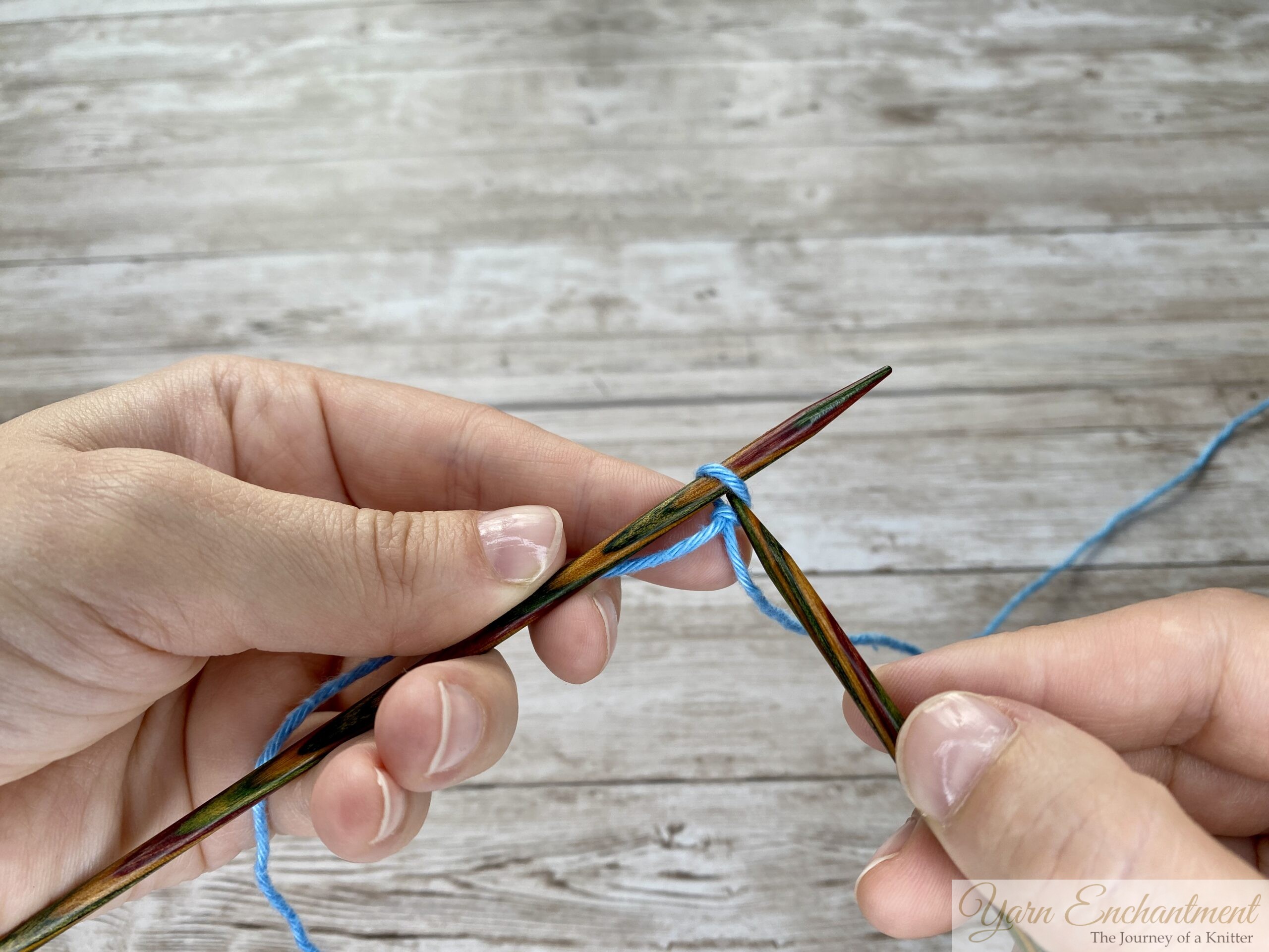Knitting needle inserted into a loop of blue yarn for the first step of a knitted cast-on.