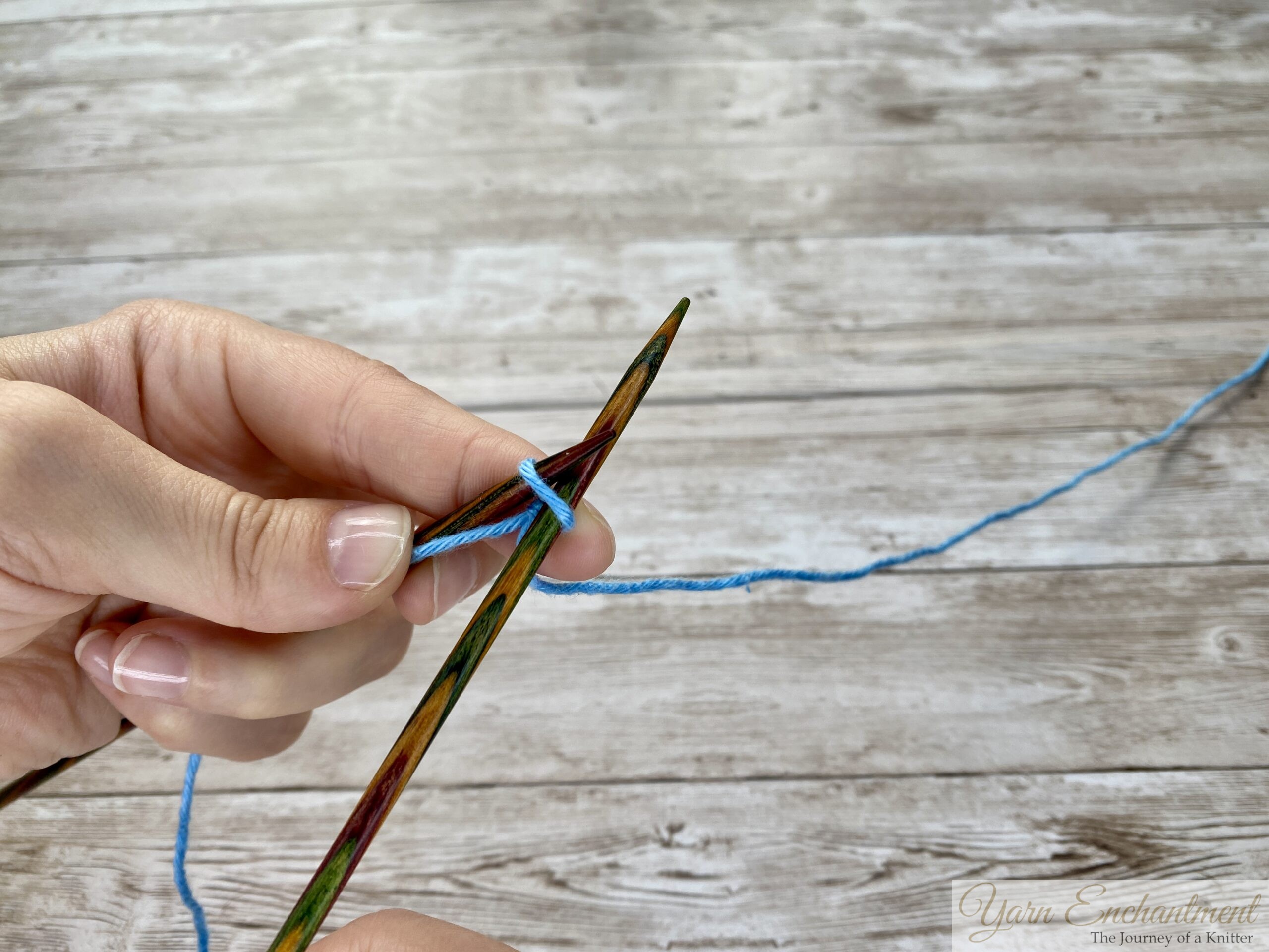 Hands maneuvering yarn and knitting needles to create a new loop during a knitted cast-on.