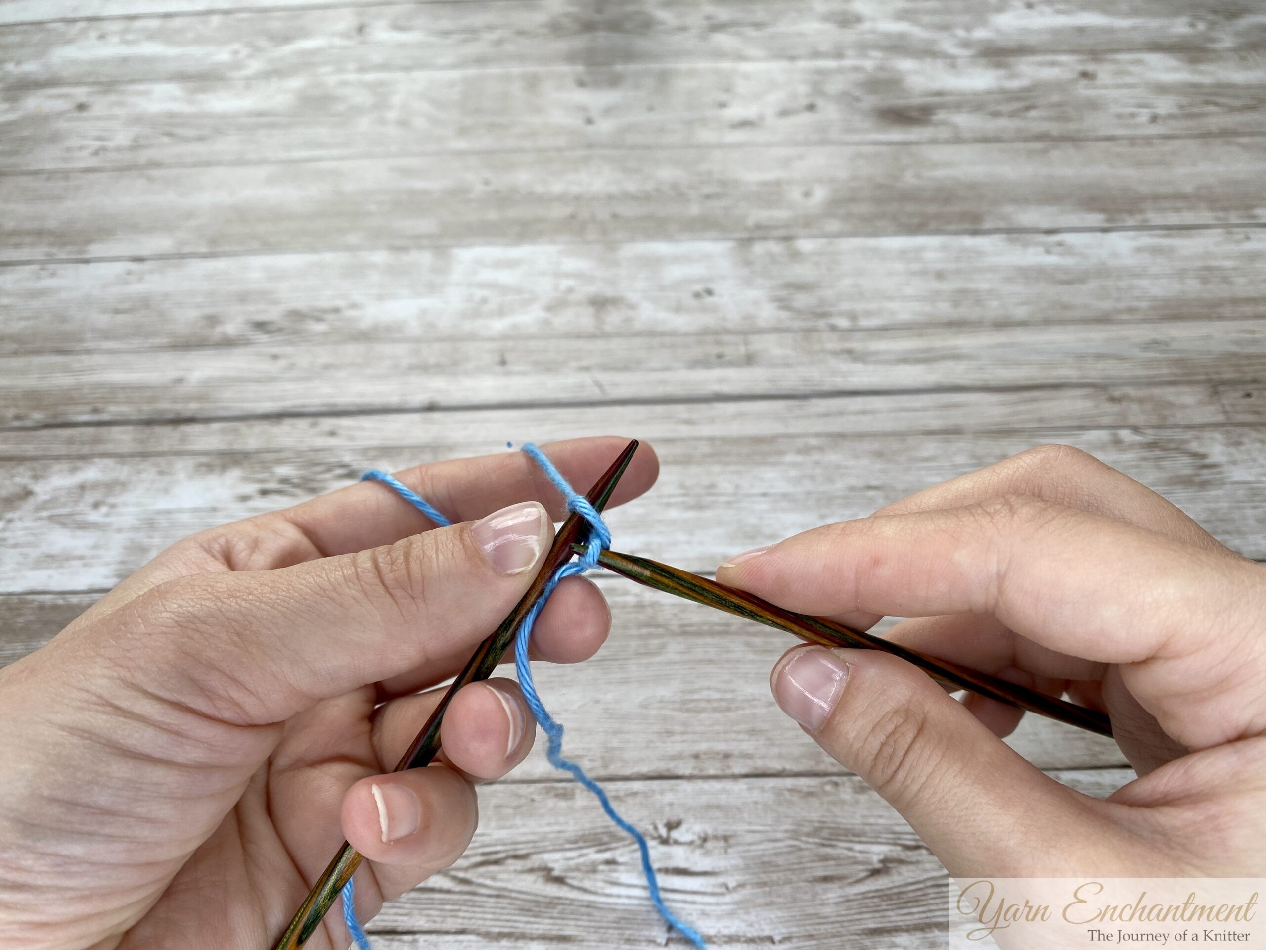 Hands inserting the right needle into a loop on the left needle during a knitted cast-on.
