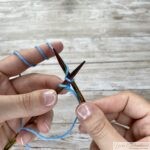 Hands positioning knitting needles and blue yarn to pull through a loop during a knitted cast-on.