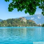 Scenic view of a turquoise lake surrounded by lush green hills and forested mountains under a clear blue sky. In the center background, a historic castle sits atop a rocky hill, and a church with a tall steeple is visible to the right. Overhanging green tree branches frame the top of the image, enhancing the vibrant, summery atmosphere.