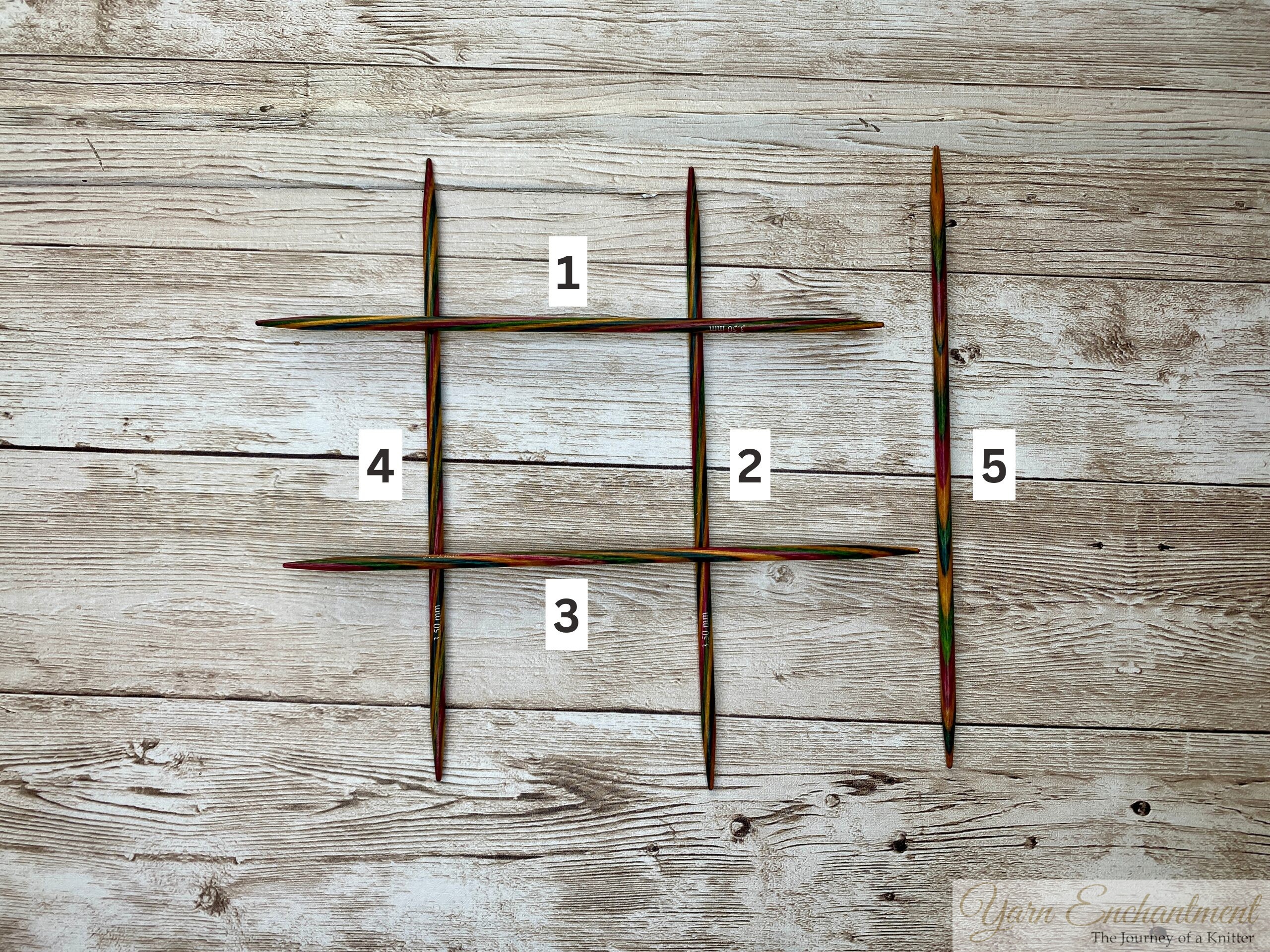 Five colorful wooden knitting needles are arranged on a light wooden surface. Four of the needles form a large hashtag or tic-tac-toe grid shape on the left side of the image, while the fifth needle stands vertically on the right, separate from the grid. The background is a textured, pale wood. No yarn or other objects are visible.