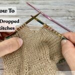 Hands holding colorful knitting needles, demonstrating how to fix a dropped stitch in beige stockinette fabric. Several horizontal yarn strands, called ladders, are visible above the dropped stitch.