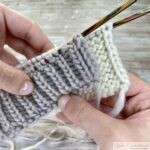 A close-up of hands holding a piece of white knitting on wooden needles, showing a swatch of grey brioche stitch and white garter stitch with a clear view of the neat selvedge edge, demonstrating the edge technique on a rustic wooden background.