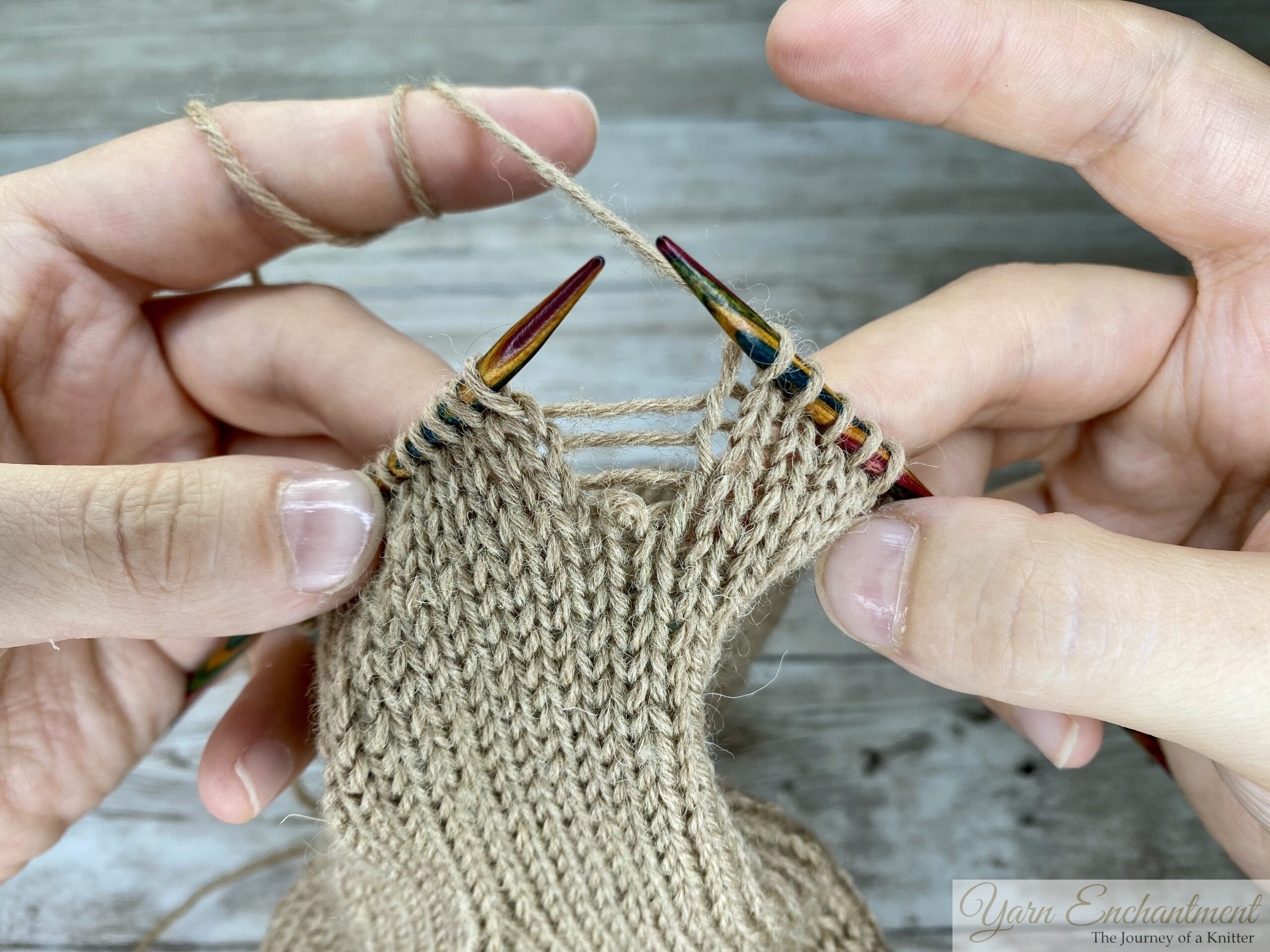 Hands holding knitting needles and yarn, demonstrating how to fix a dropped stitch in beige stockinette fabric. Several horizontal yarn strands, known as ladders, are visible where the stitch has unraveled.