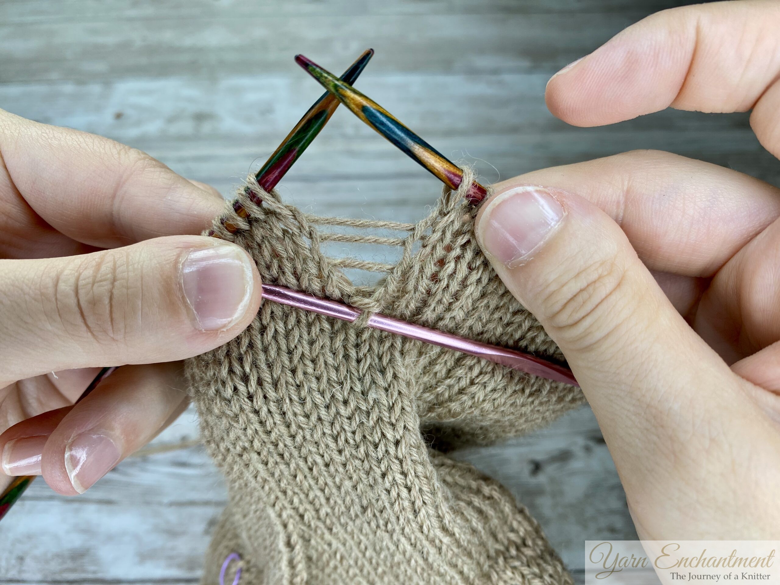 Hands holding colorful double-pointed knitting needles and a crochet hook, preparing to fix a dropped stitch in beige stockinette fabric. Several horizontal yarn strands (ladders) are visible above the dropped stitch.