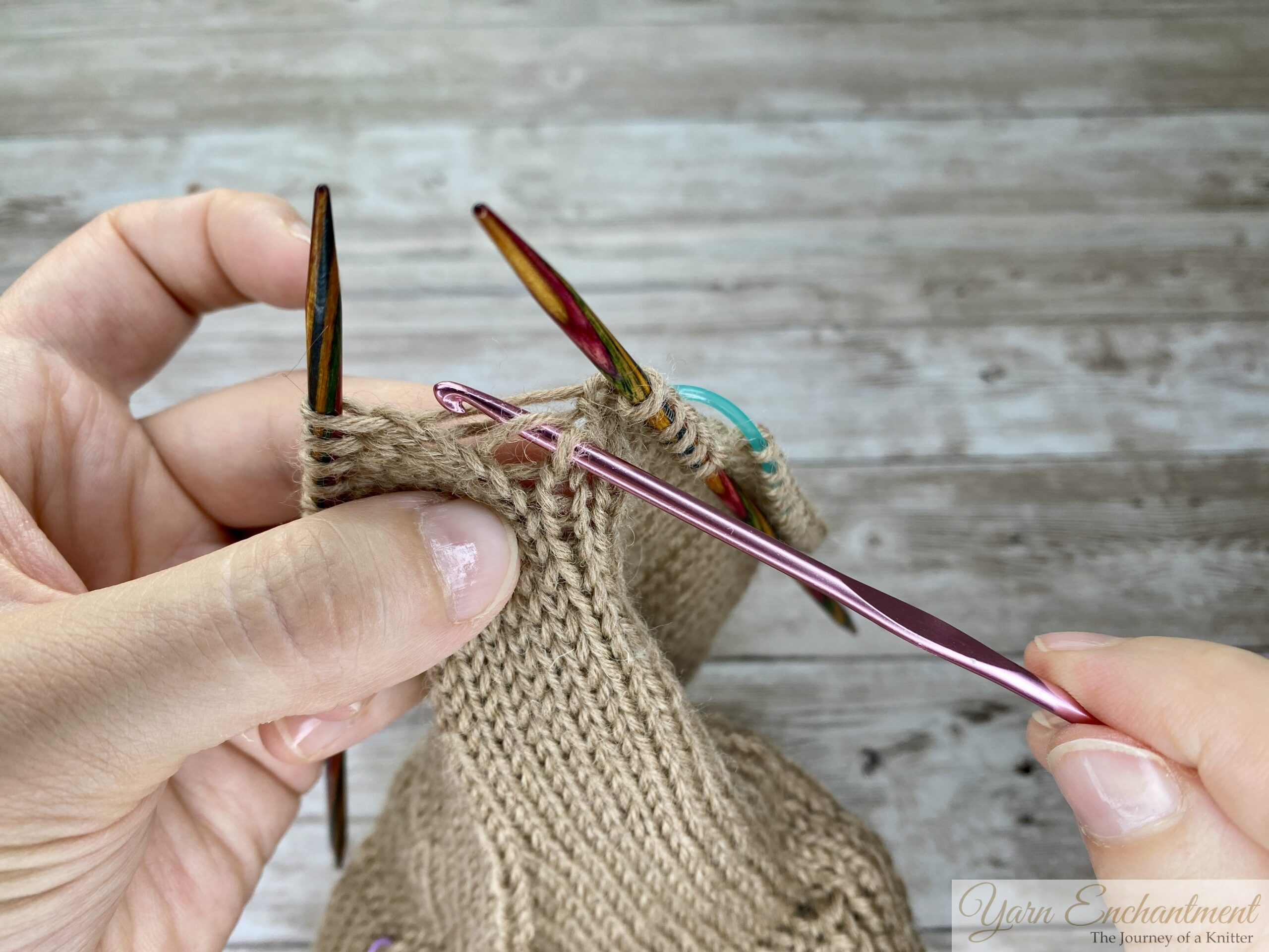 Close-up of hands inserting a crochet hook under the first horizontal ladder strand to pick up a dropped stitch in beige stockinette knitting, with colorful needles holding the surrounding stitches.