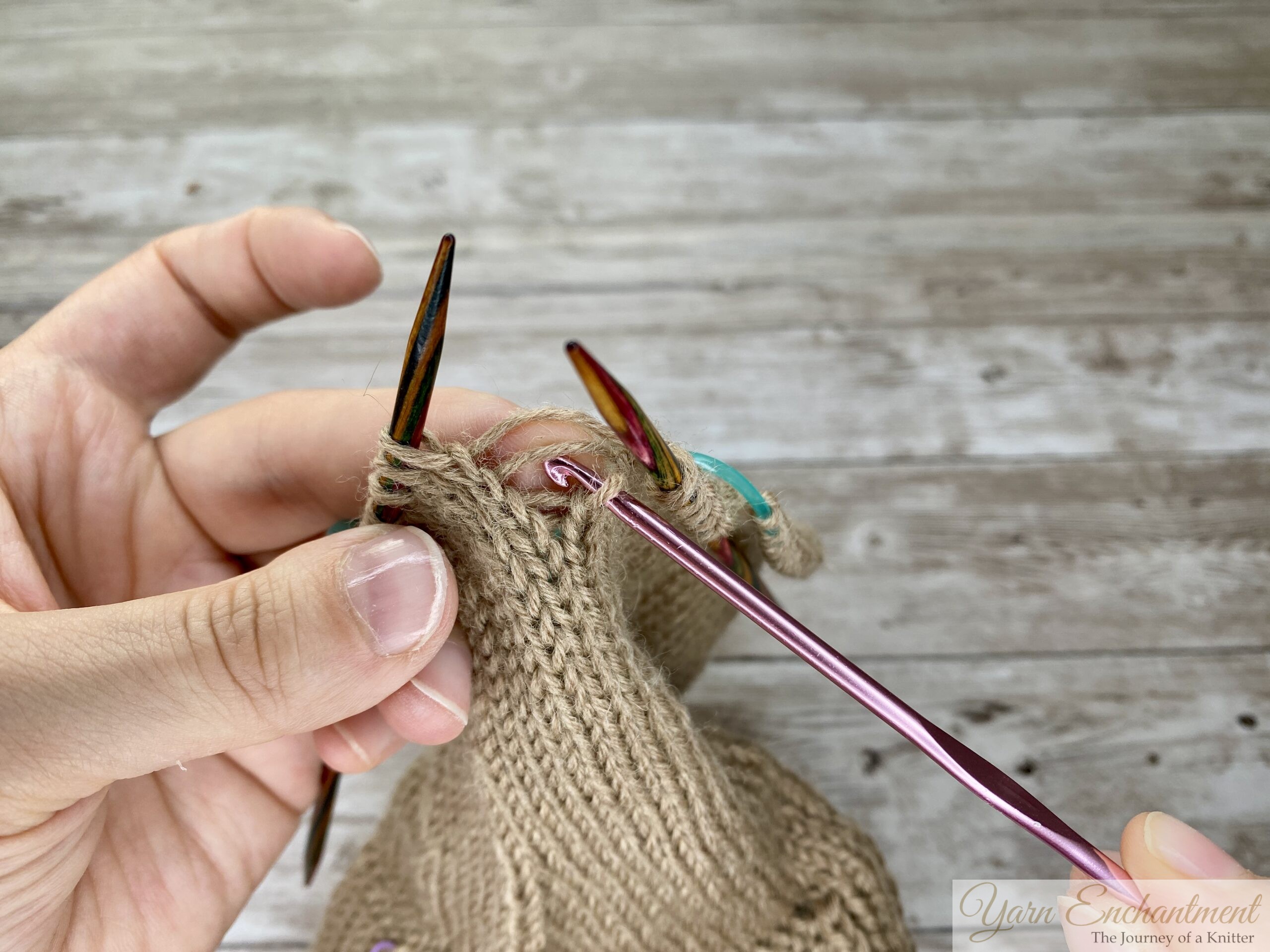 Close-up of hands inserting a crochet hook under the first horizontal ladder strand to pick up a dropped stitch in beige stockinette knitting, with colorful needles holding the surrounding stitches.